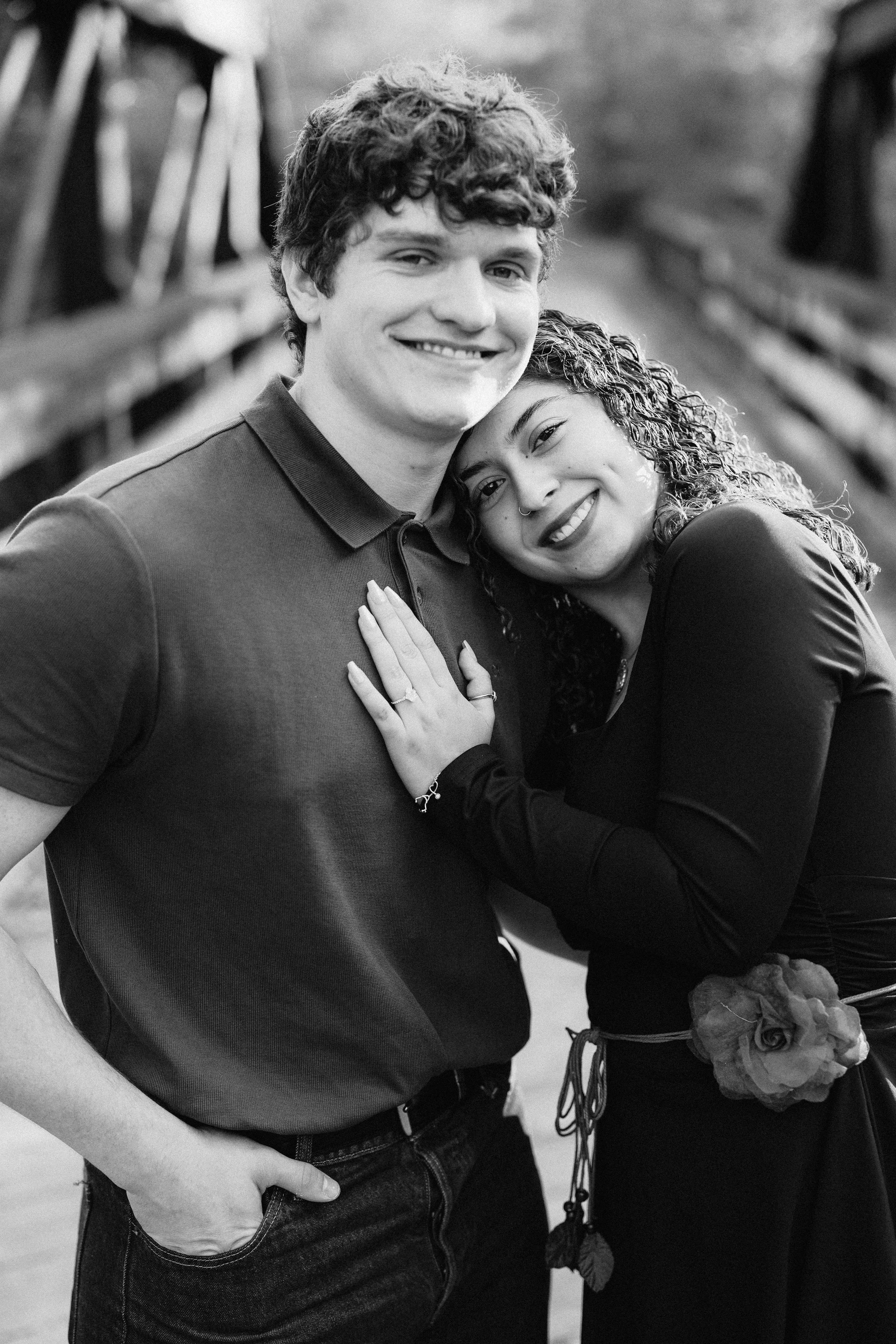 A smiling young man and woman hugging each other, with the woman resting her head on the man’s shoulder, on a bridge outdoors.
