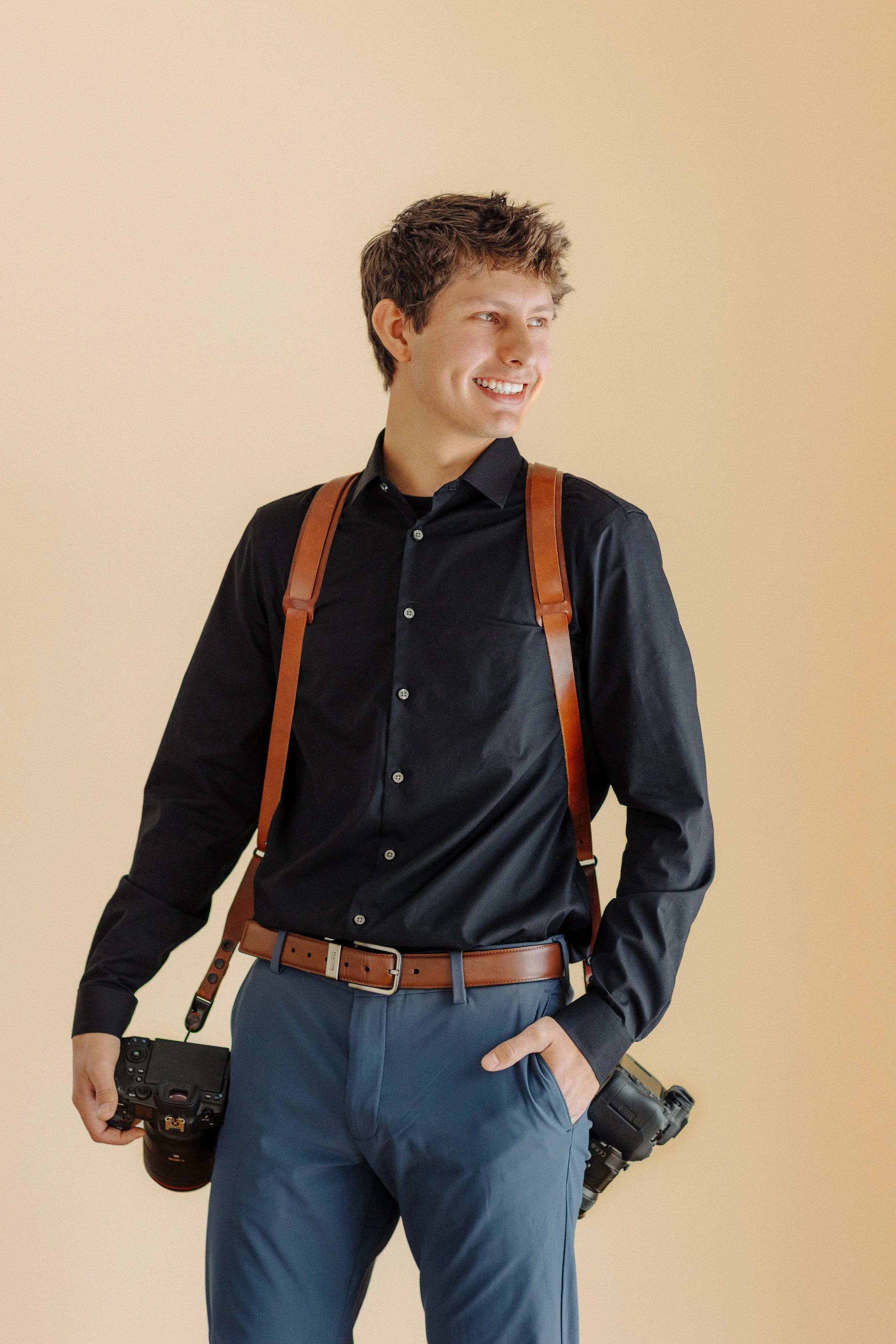 A young man in a black shirt and blue pants with a brown camera strap, holding a camera in his right hand, smiling and looking to his right, against a plain beige background.