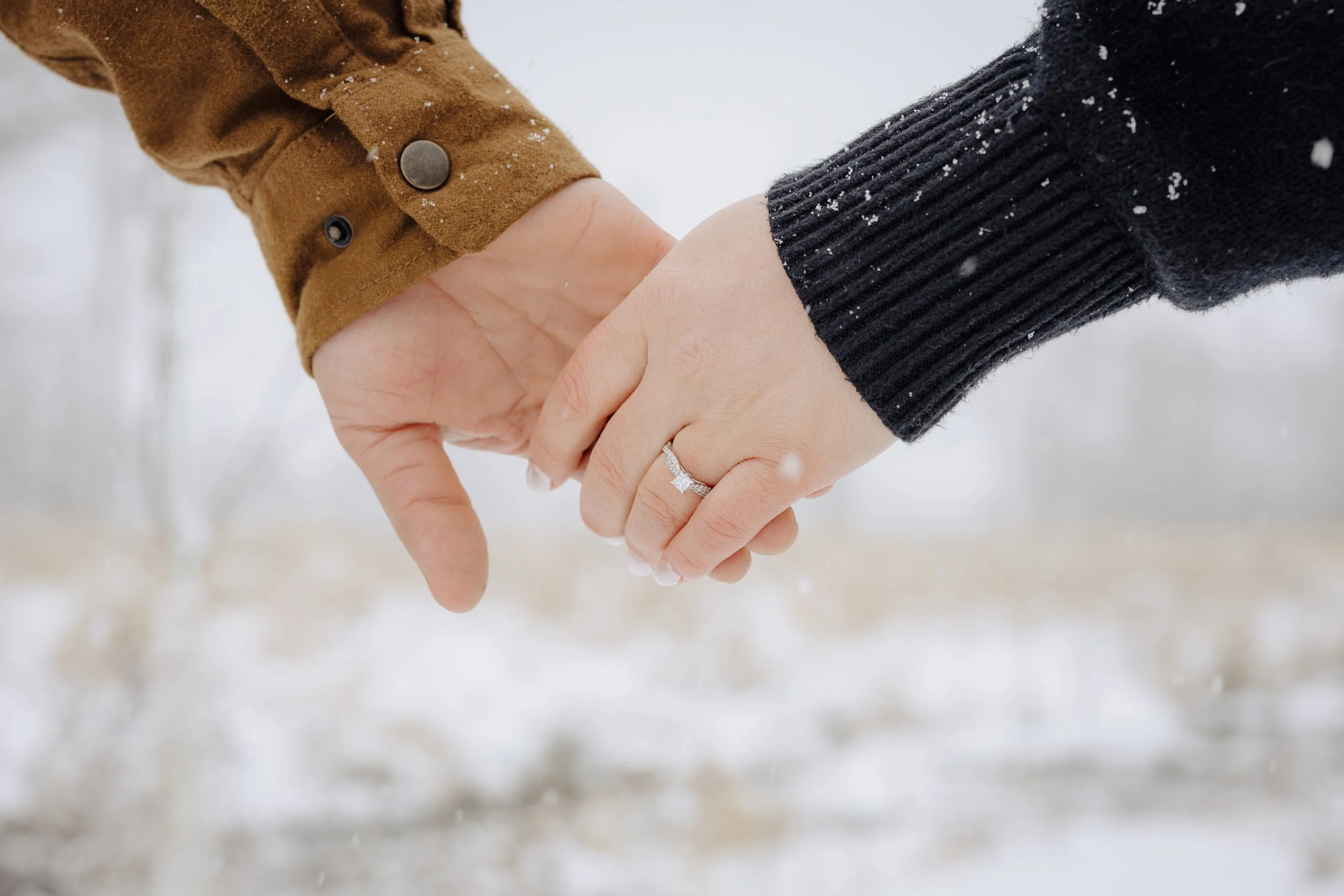 Close-up of two people holding hands in the snow, one wearing a black sweater and the other in a brown jacket, with a ring on the woman's finger.