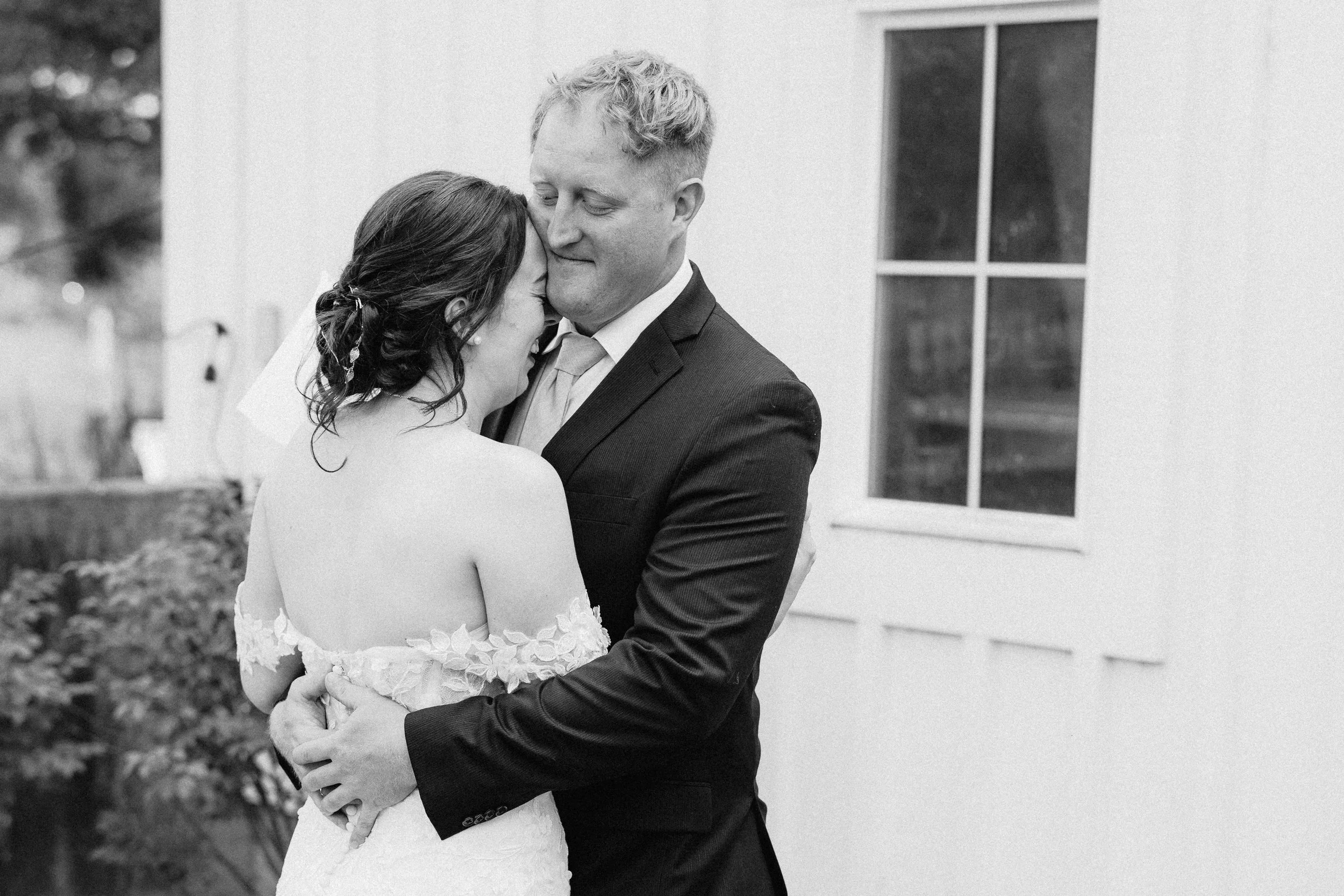 Black and white photograph of a bride and groom embracing each other, with the bride resting her head on the groom's chest and the groom gently holding her. They are standing outside near a white building with a window in the background.