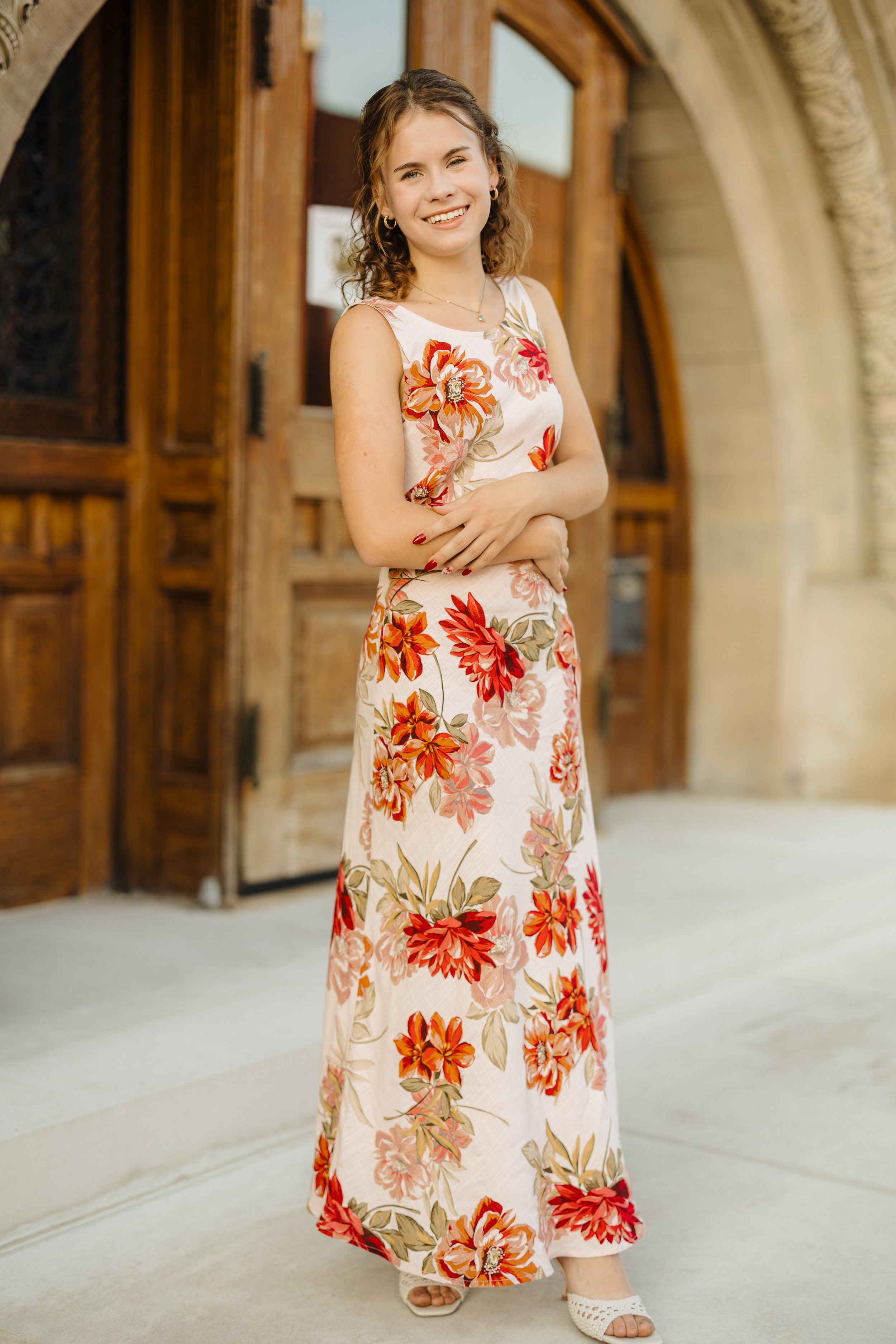 Young woman with curly hair smiling and standing outdoors in front of wooden doors, wearing a sleeveless floral maxi dress and jewelry.