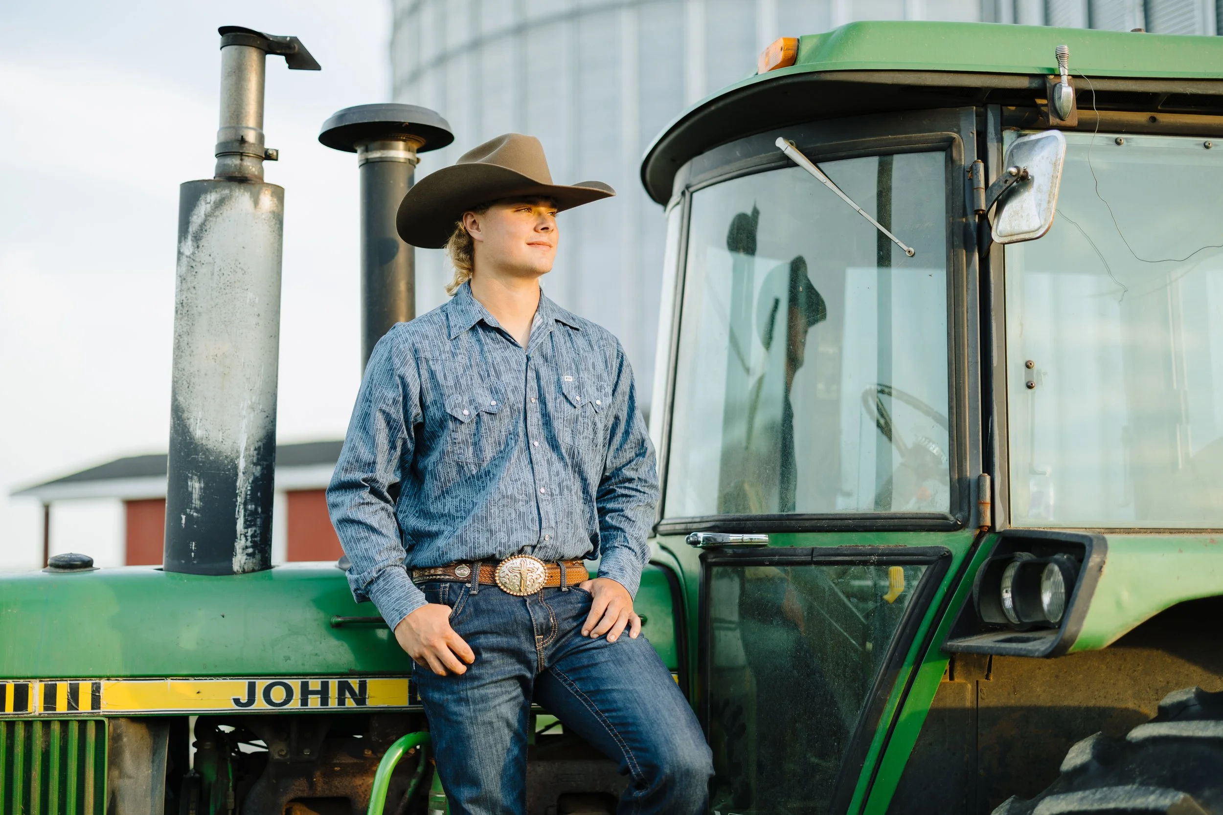 A man wearing a cowboy hat, denim shirt, and jeans leaning against a green John Deere tractor outside near a barn.