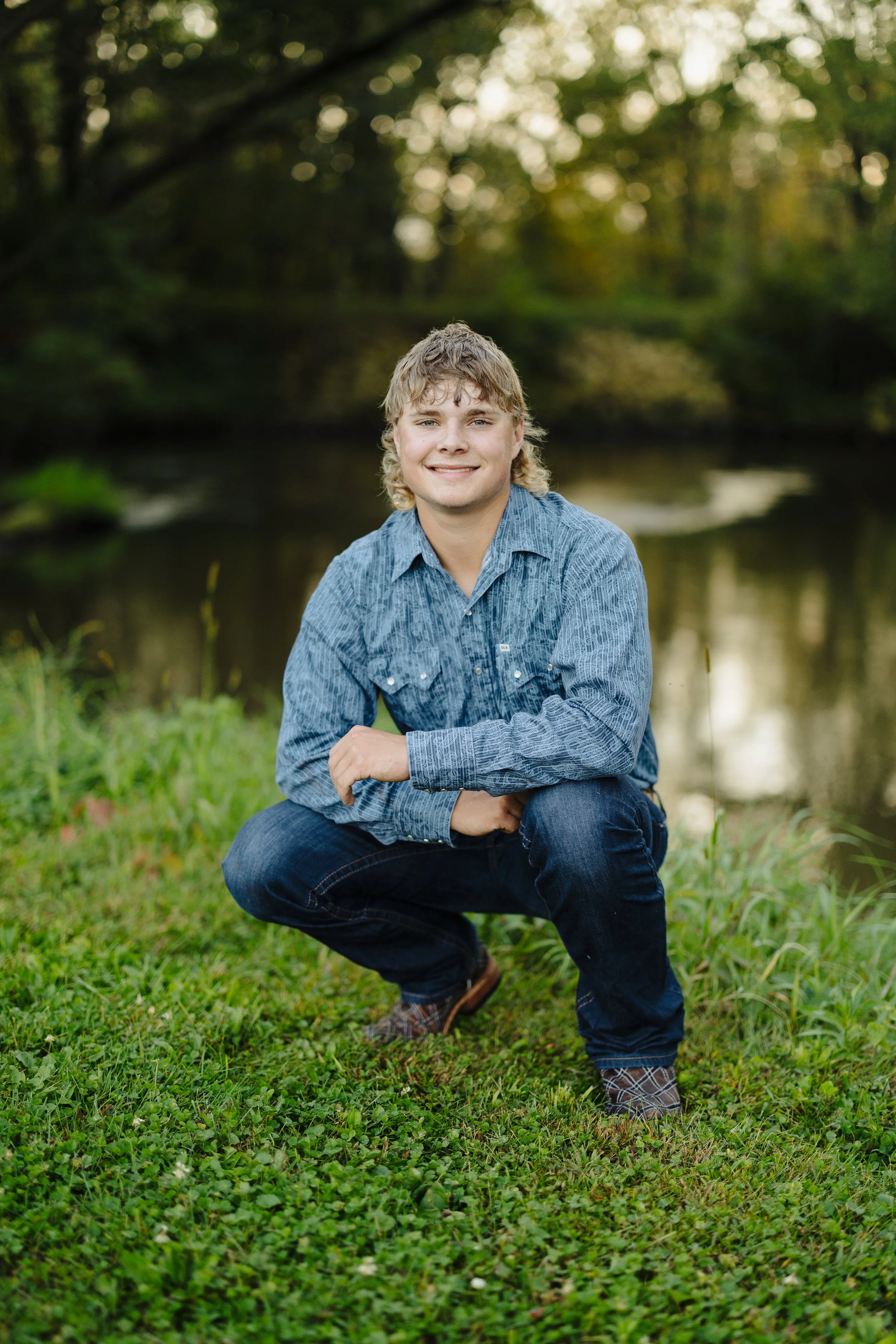 Young man kneeling on grass near a river, wearing a blue shirt and jeans, smiling at the camera with trees in the background.