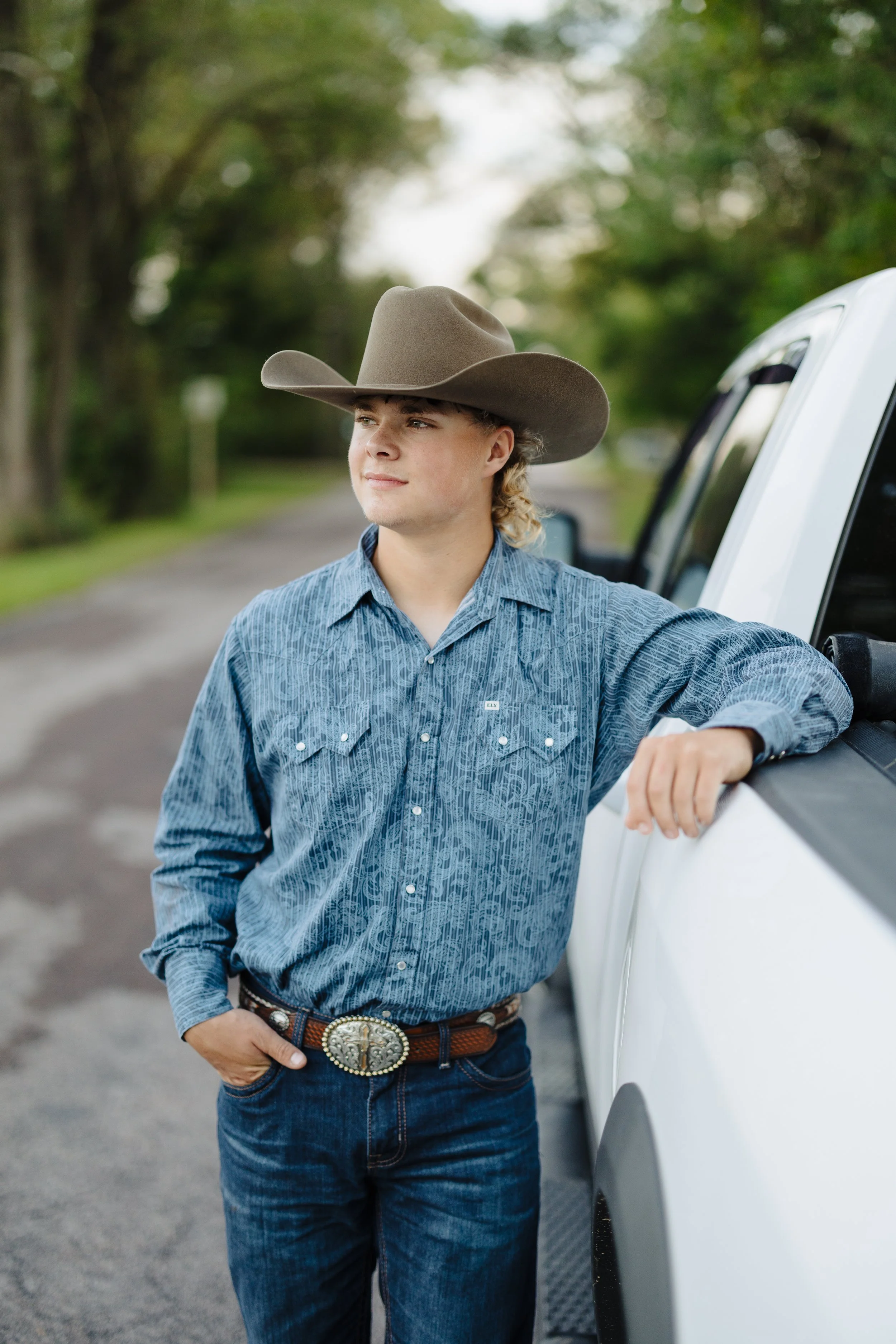 Young man dressed in cowboy attire, wearing a large cowboy hat, a patterned blue shirt, and jeans, leaning on a white truck with one hand resting on the door, outdoors in a rural setting.