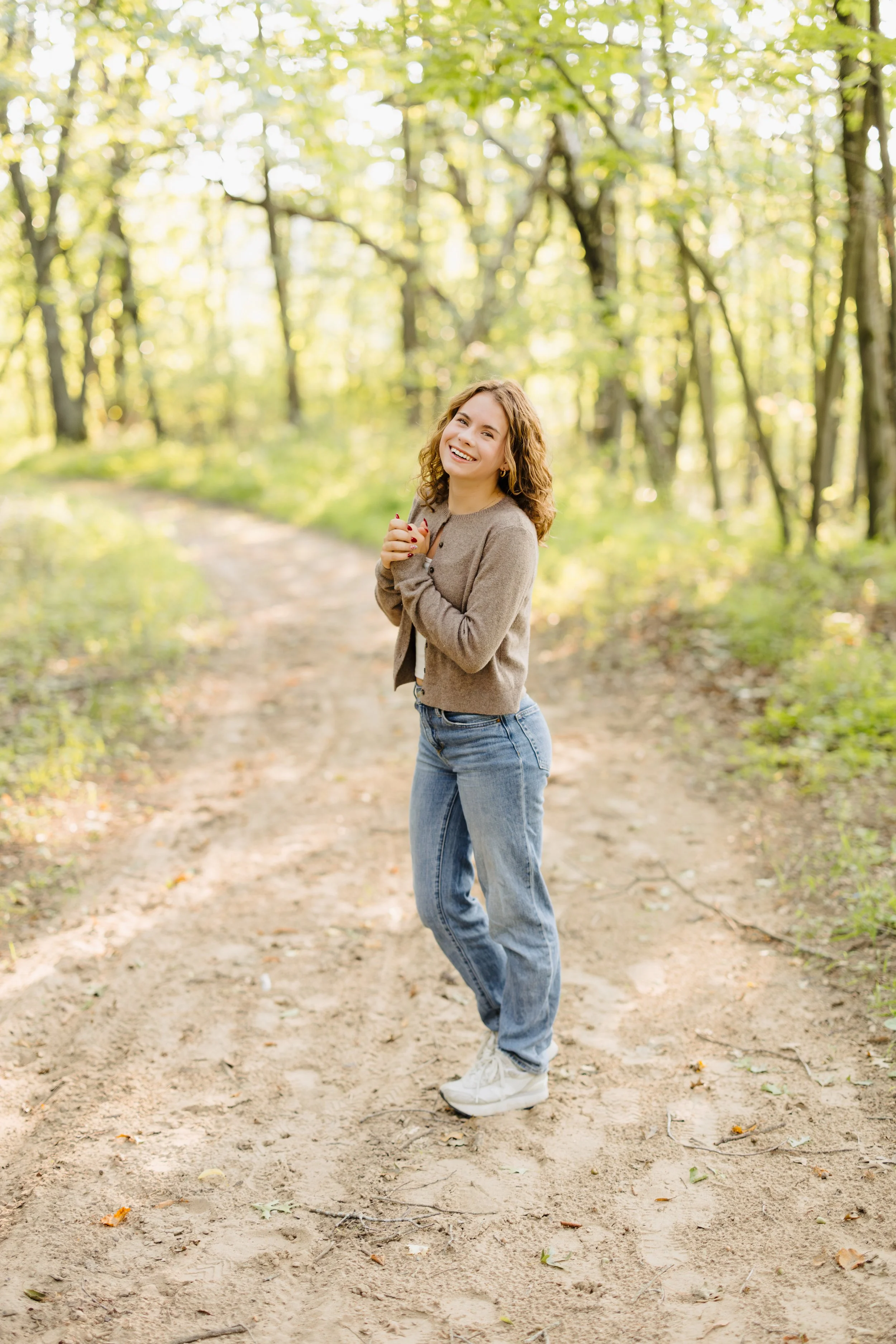 A young woman smiling and standing on a dirt path in a lush green forest during daytime.