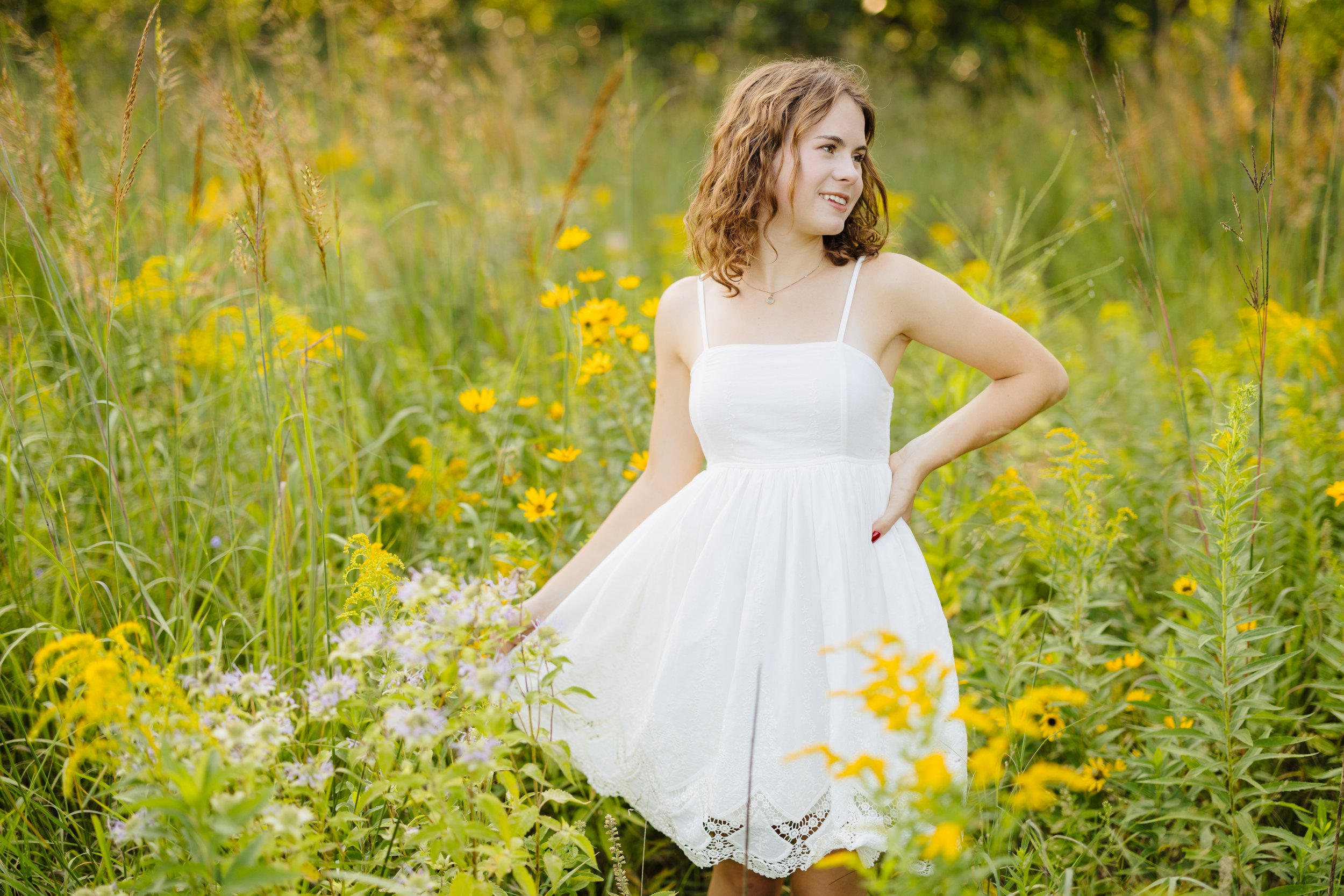 A young woman in a white dress standing in a field of yellow wildflowers and tall grass, with trees in the background, smiling and looking to the side.