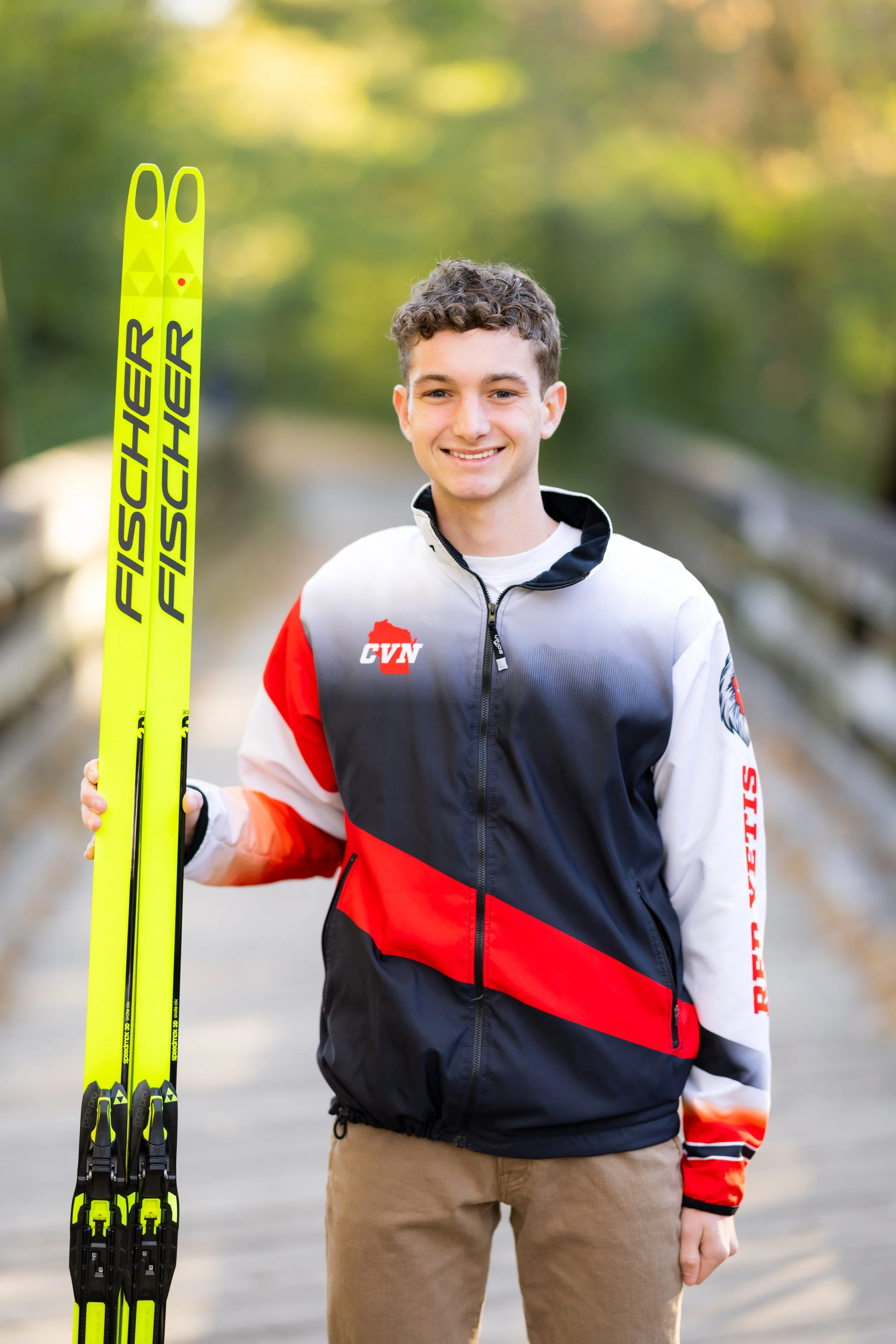 A young man smiling and holding a pair of yellow Fischer cross-country skis outdoors on a sunny day.