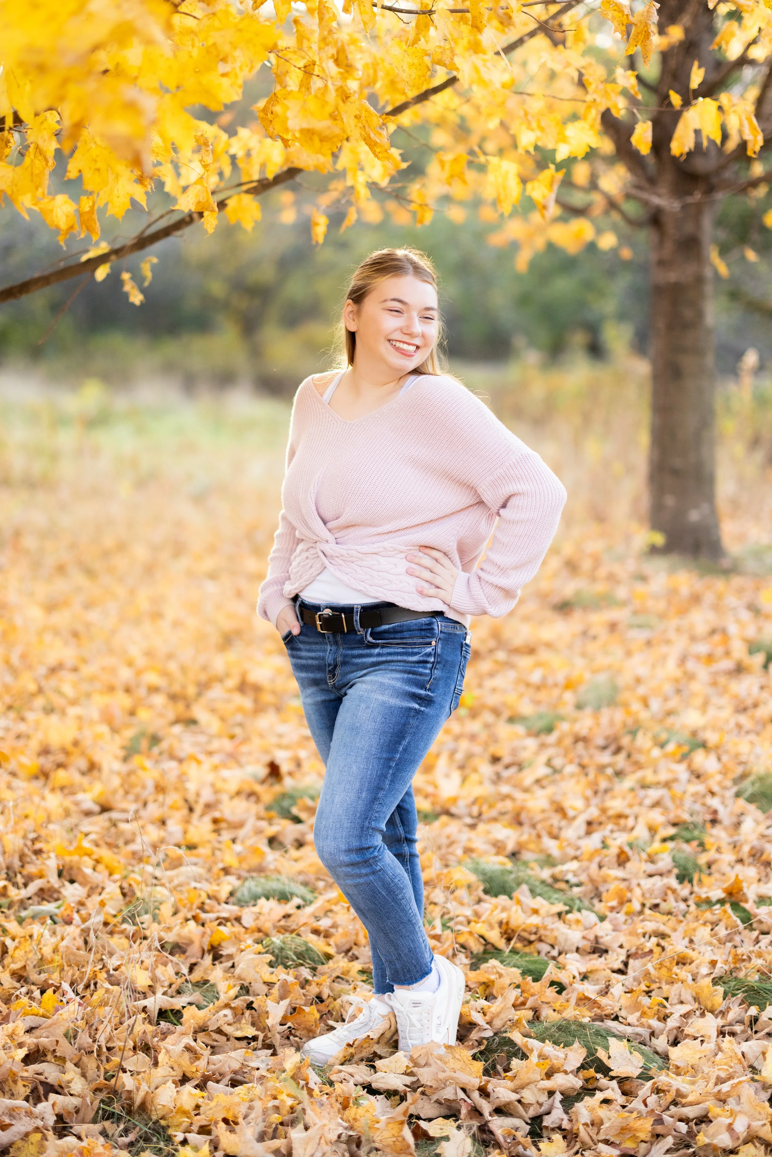 A young woman in a pink sweater, jeans, and white sneakers standing outdoors on a bed of fallen autumn leaves, smiling and looking to her right, with a large tree with yellow leaves behind her.