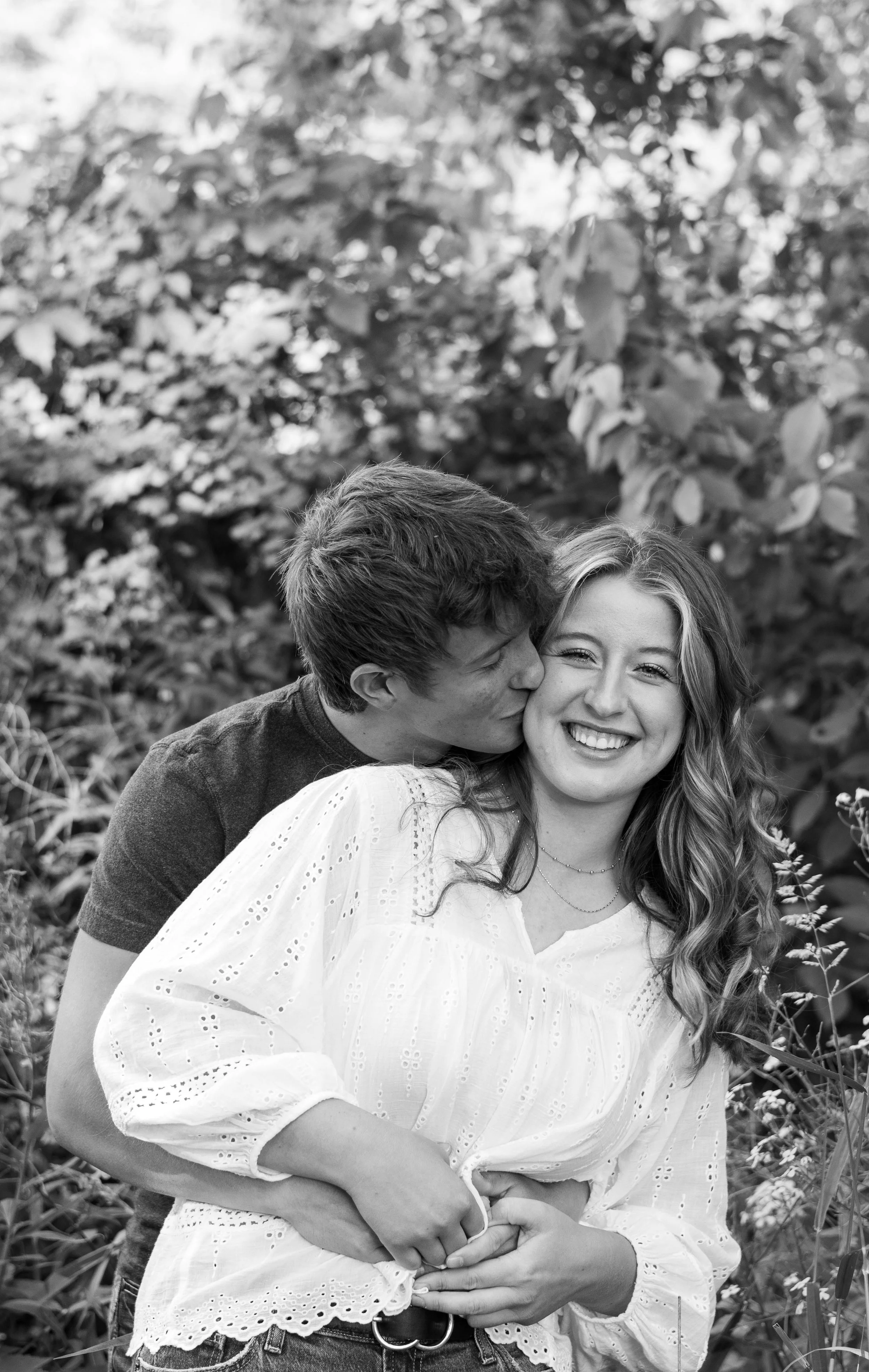 A smiling young woman with wavy hair standing outdoors with a person kissing her on the cheek, surrounded by foliage in a natural setting.