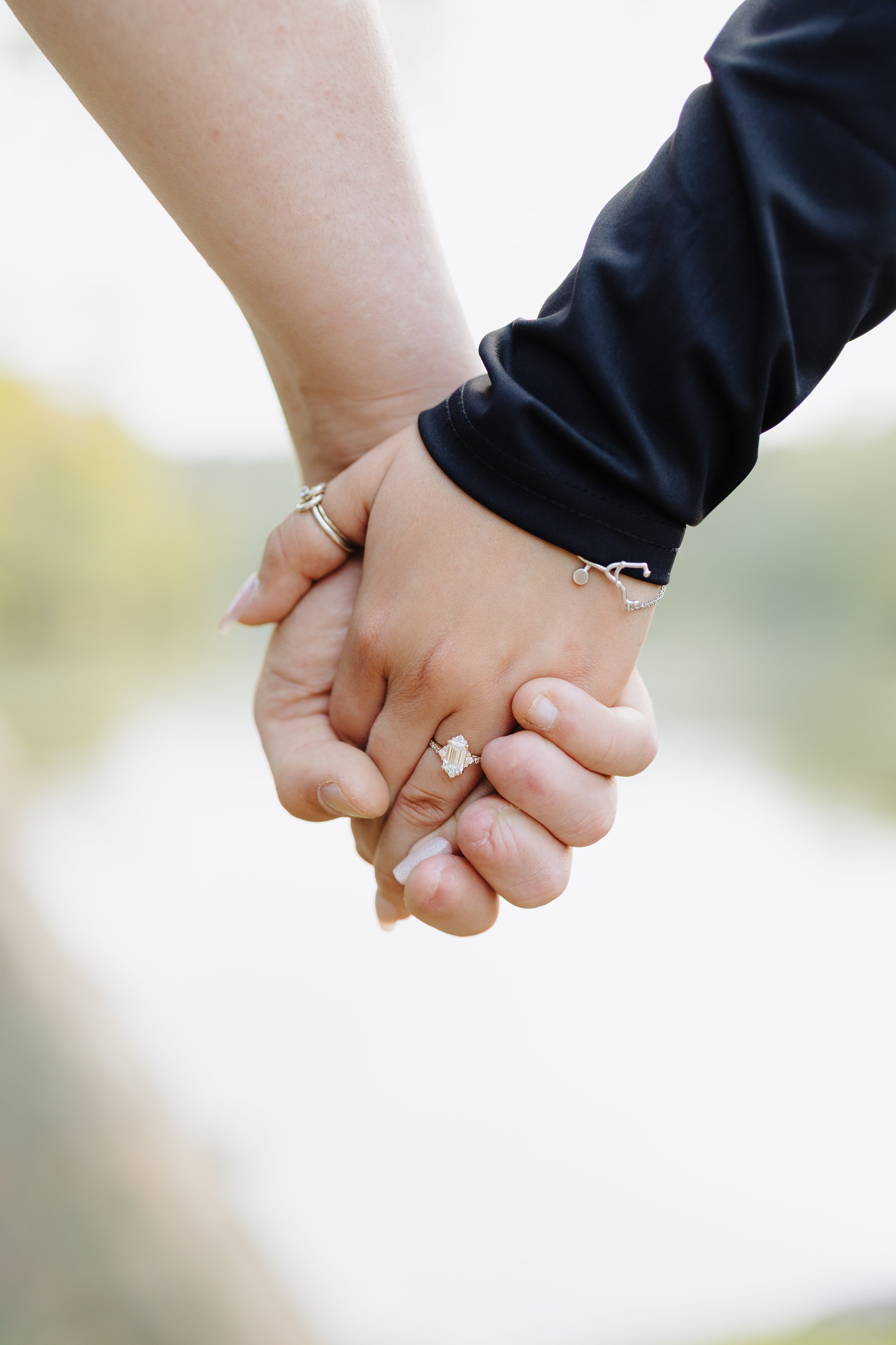 Close-up of two people holding hands, with visible jewelry including rings and bracelets, against a blurred outdoor background.