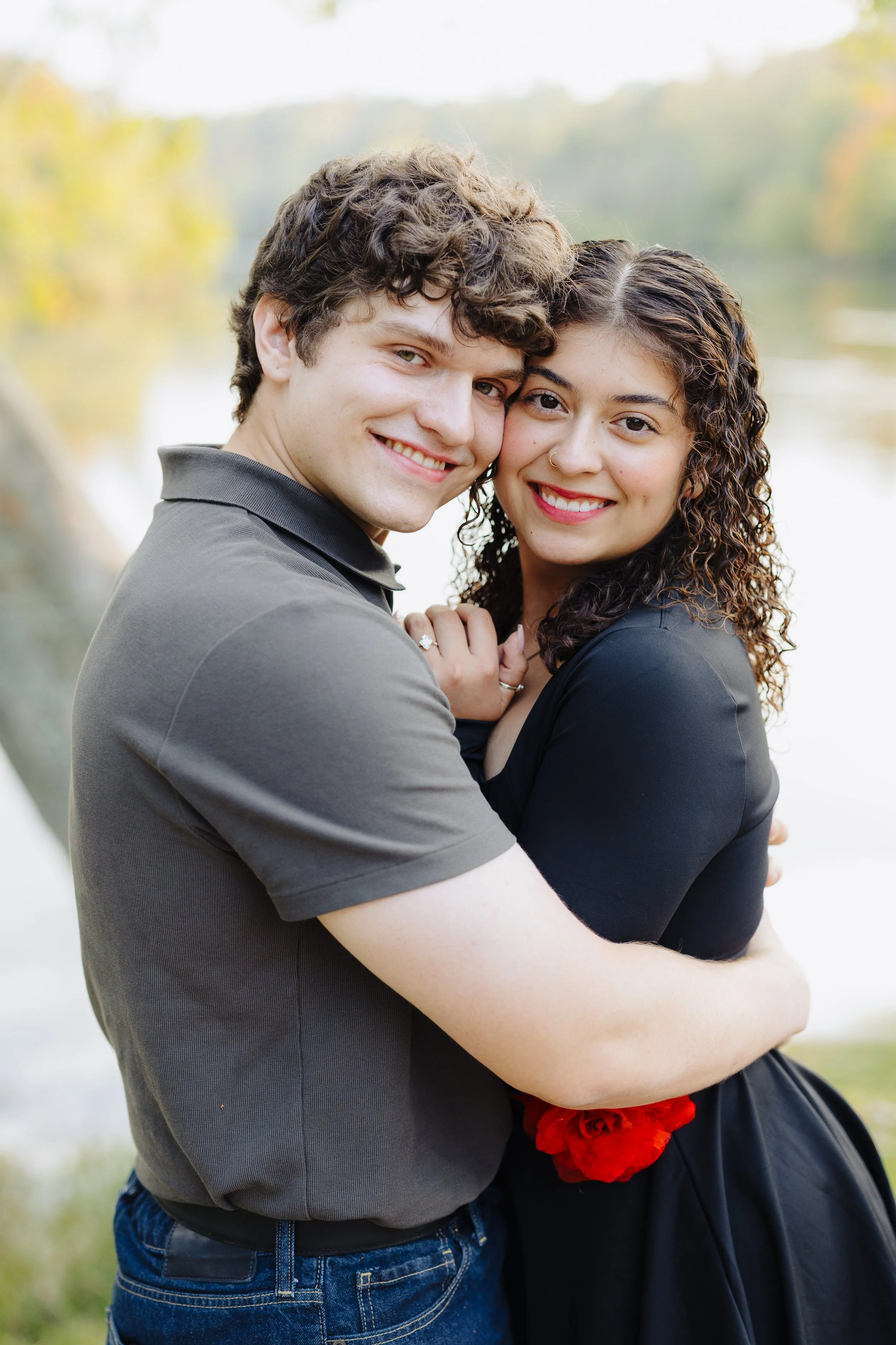 A happy couple hugging outdoors near a lake or river with autumn-colored trees in the background.