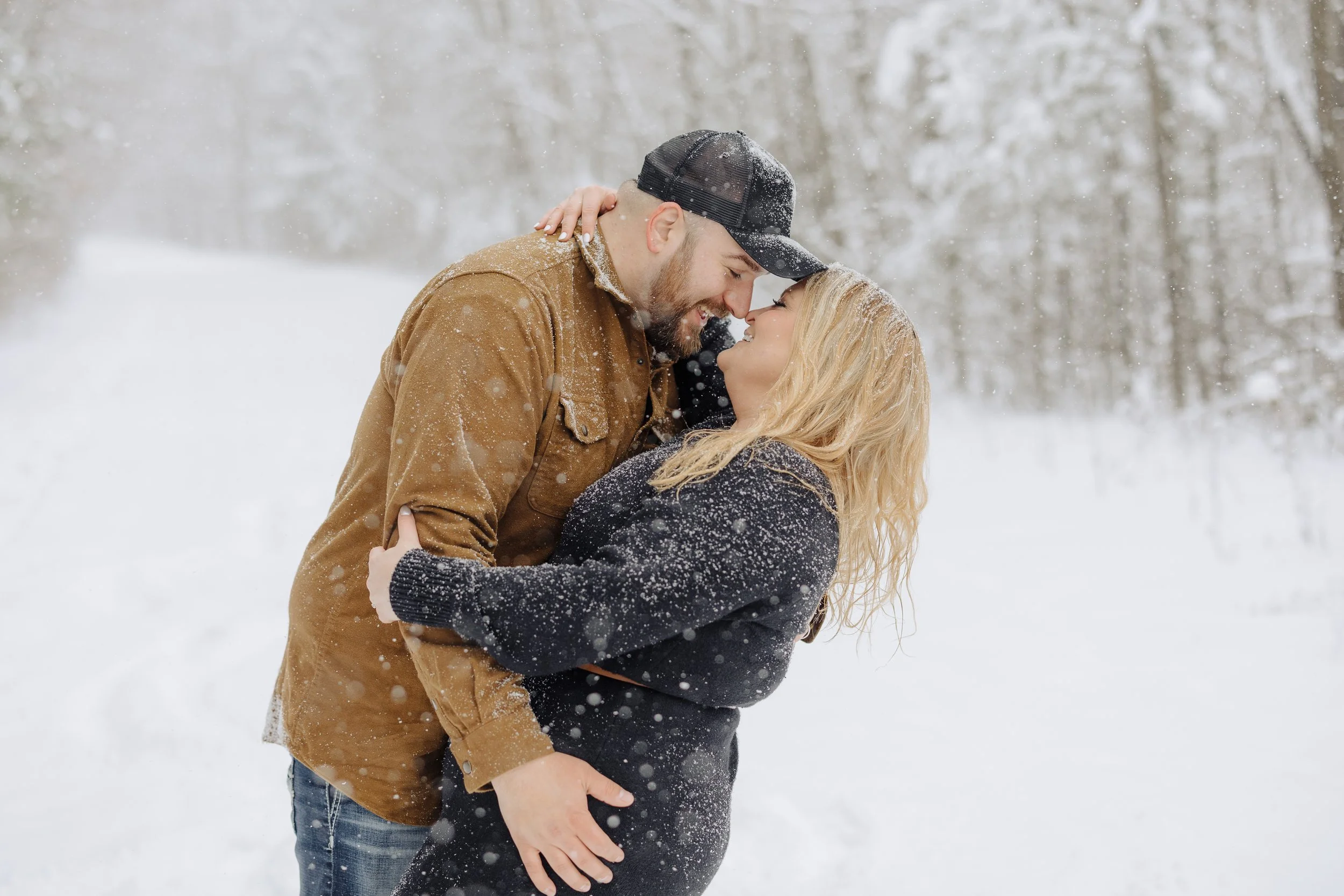 A couple sharing a loving moment in the snow, with their foreheads touching, outdoors in a snowy forest.