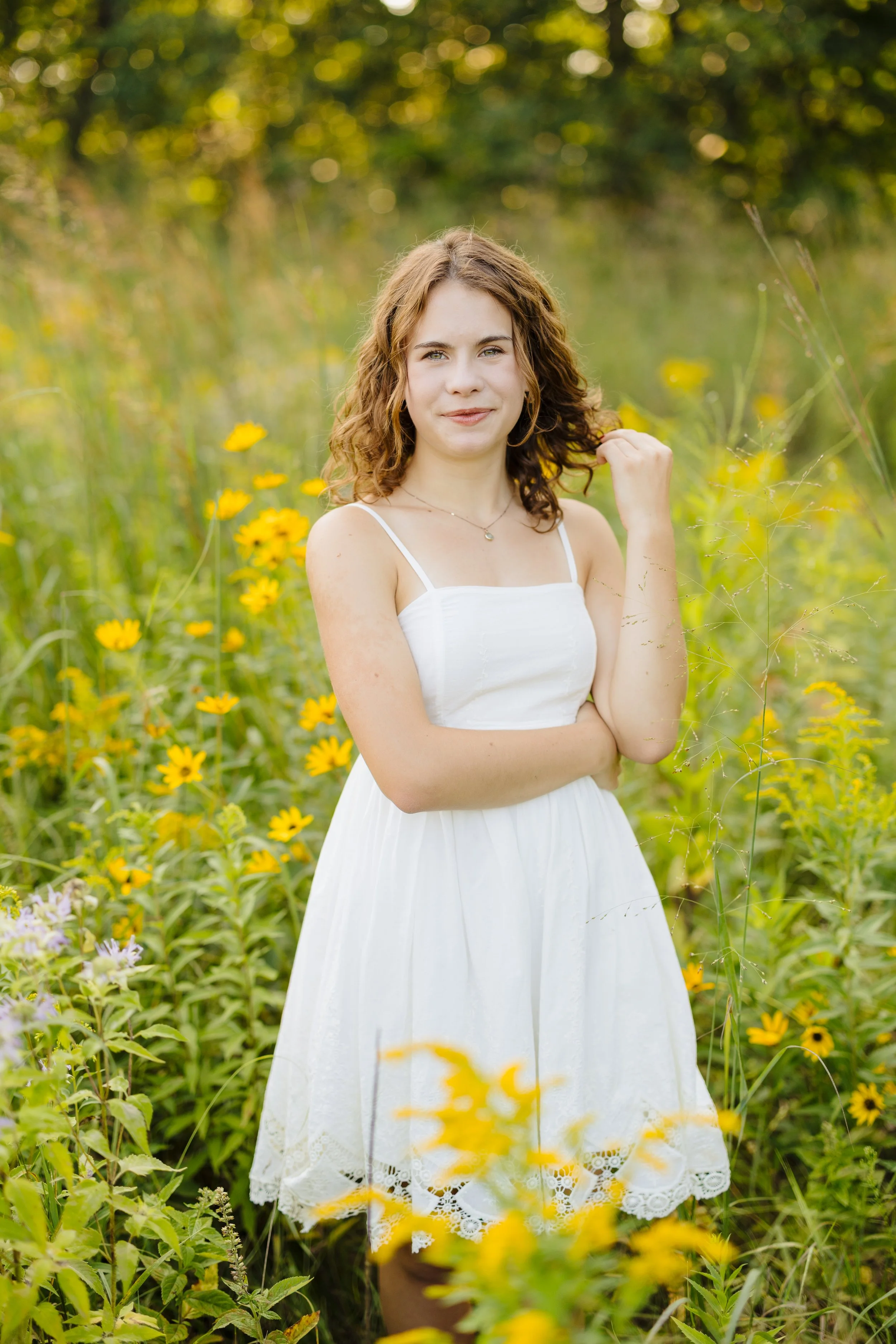 A young woman in a white dress standing in a field of yellow flowers with green foliage and trees in the background.