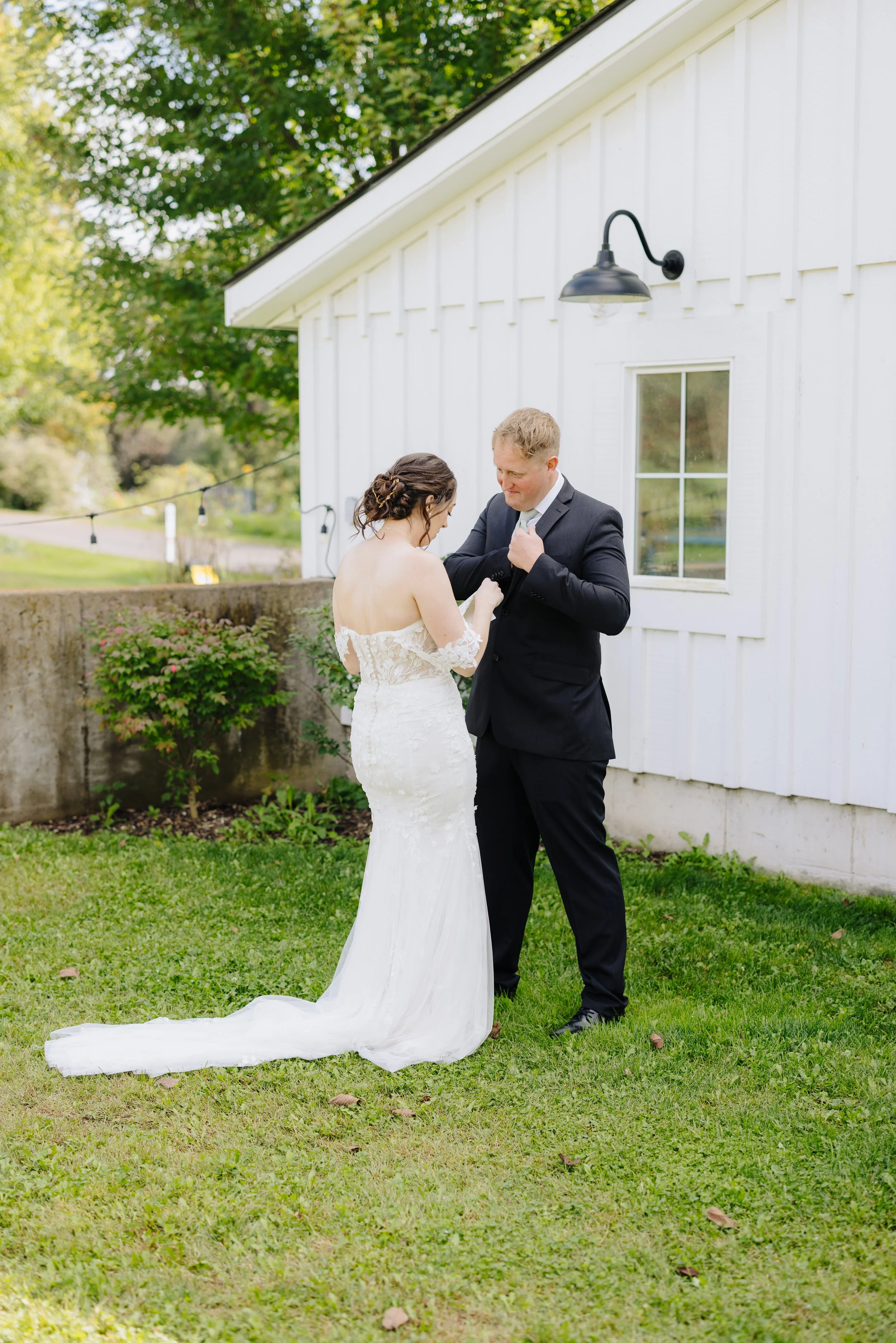 A bride and groom standing outside on a grassy area, with the bride in a white lace wedding dress and the groom in a black suit. They are looking at each other lovingly next to a white building with a black outdoor wall lamp.