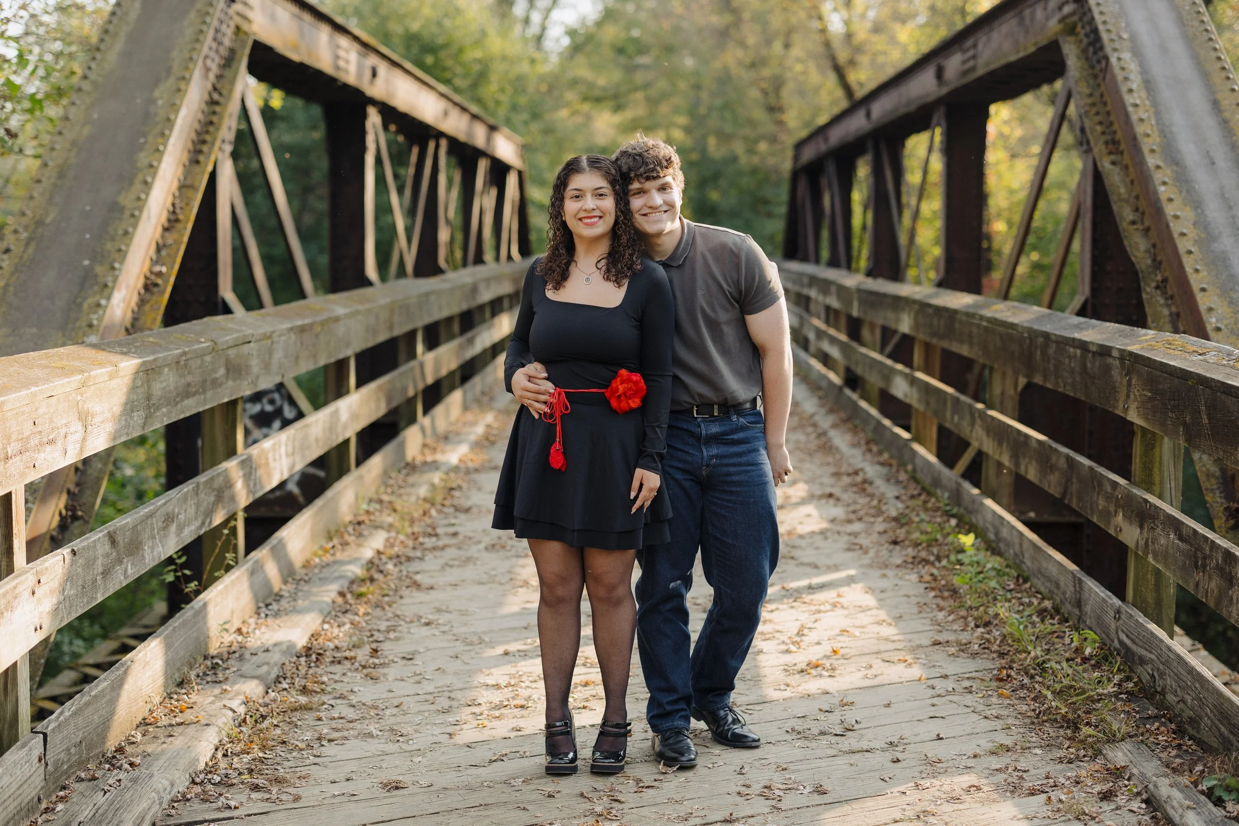 A smiling couple standing on a wooden bridge outdoors surrounded by trees with fall foliage, the woman is wearing a black dress with a red flower and a red sash, and the man is dressed in a gray polo shirt and jeans.