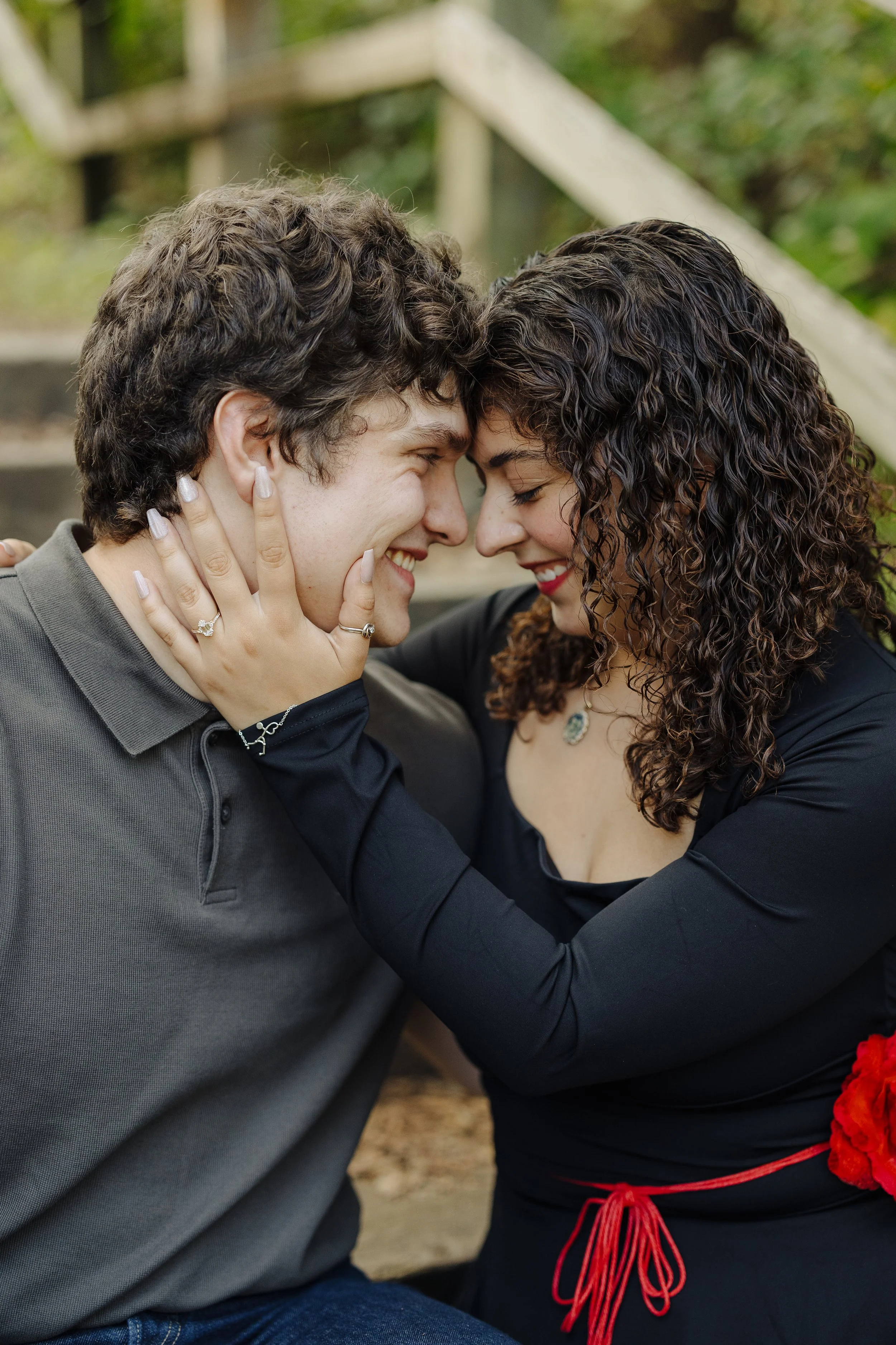 A loving couple touching foreheads and smiling, sitting outdoors near a staircase, with the woman holding the man's face gently.