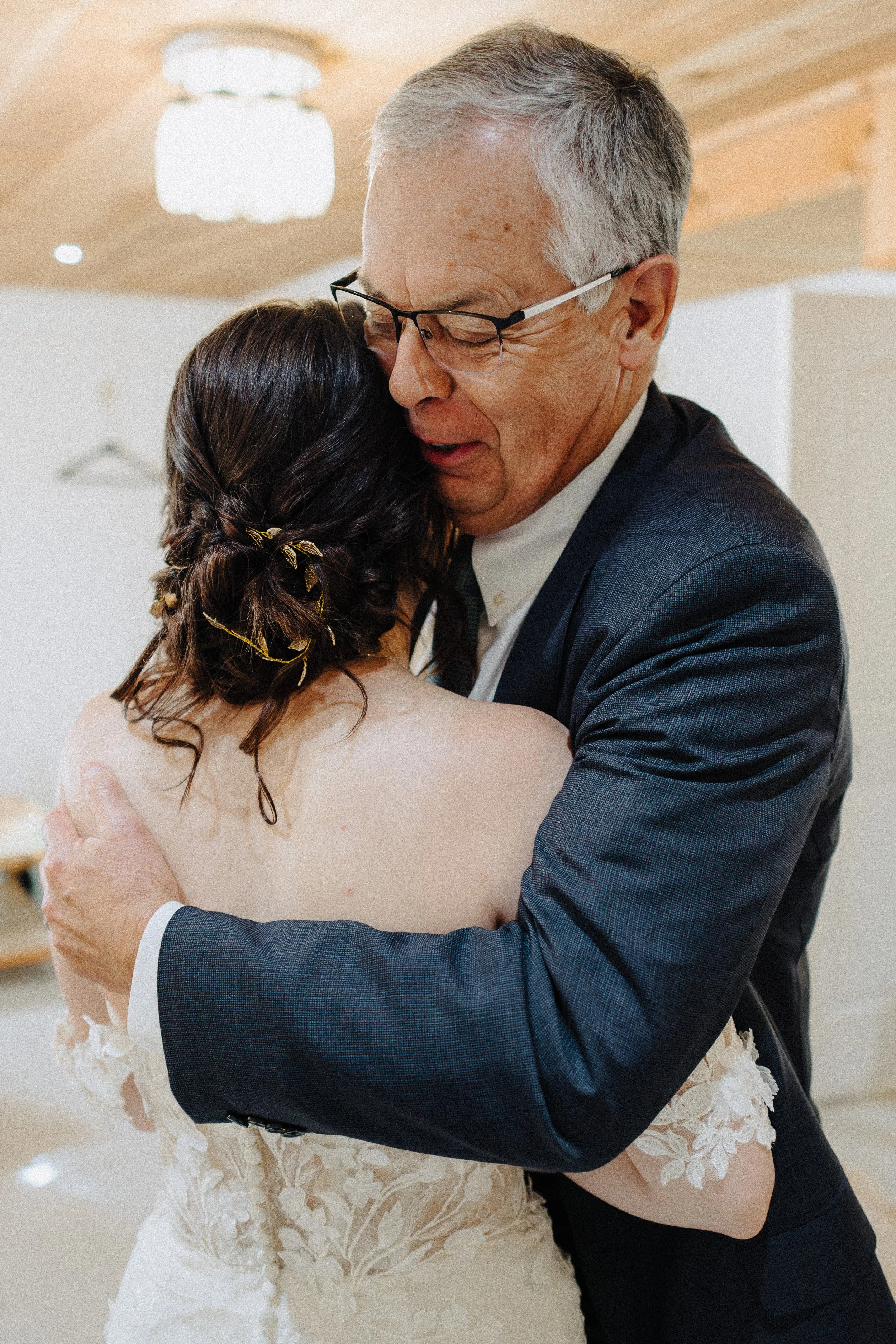 A bride and an older man, likely her father, are embracing in a heartfelt hug. The bride has dark hair styled with gold accents and wears a lace wedding dress. The man wears glasses and a dark suit. The setting appears to be a wedding venue.