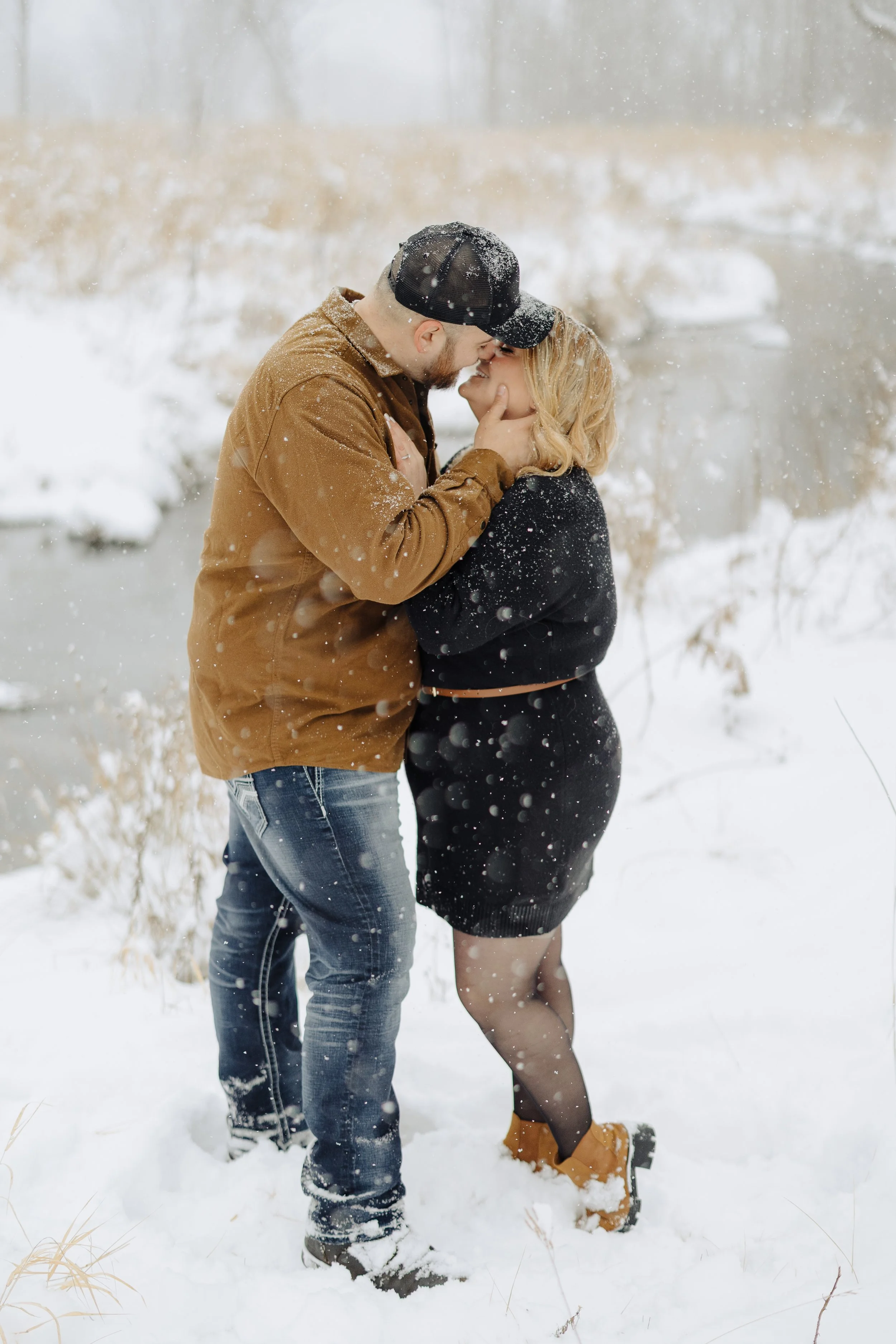 A couple sharing a kiss in a snowy outdoor setting, with the man holding the woman's face and the woman lifting her leg slightly, wearing winter clothing and snow falling around them.