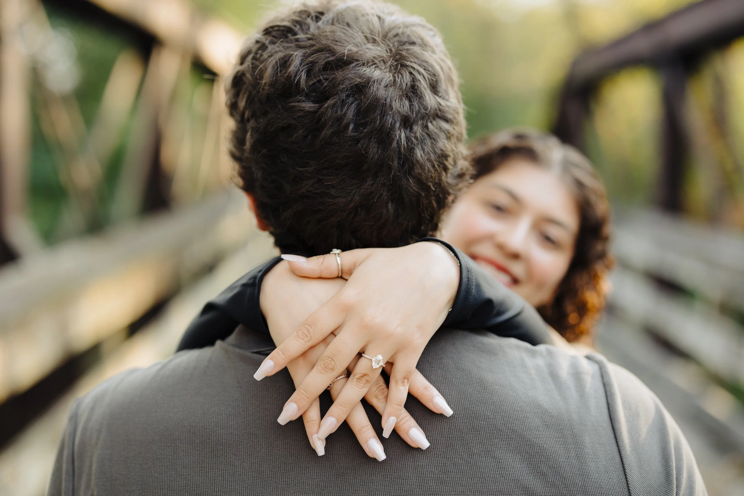 A woman with curly hair smiling at a man, her arms wrapped around his neck, showing her engagement ring. They are outdoors on a wooden bridge during daylight.