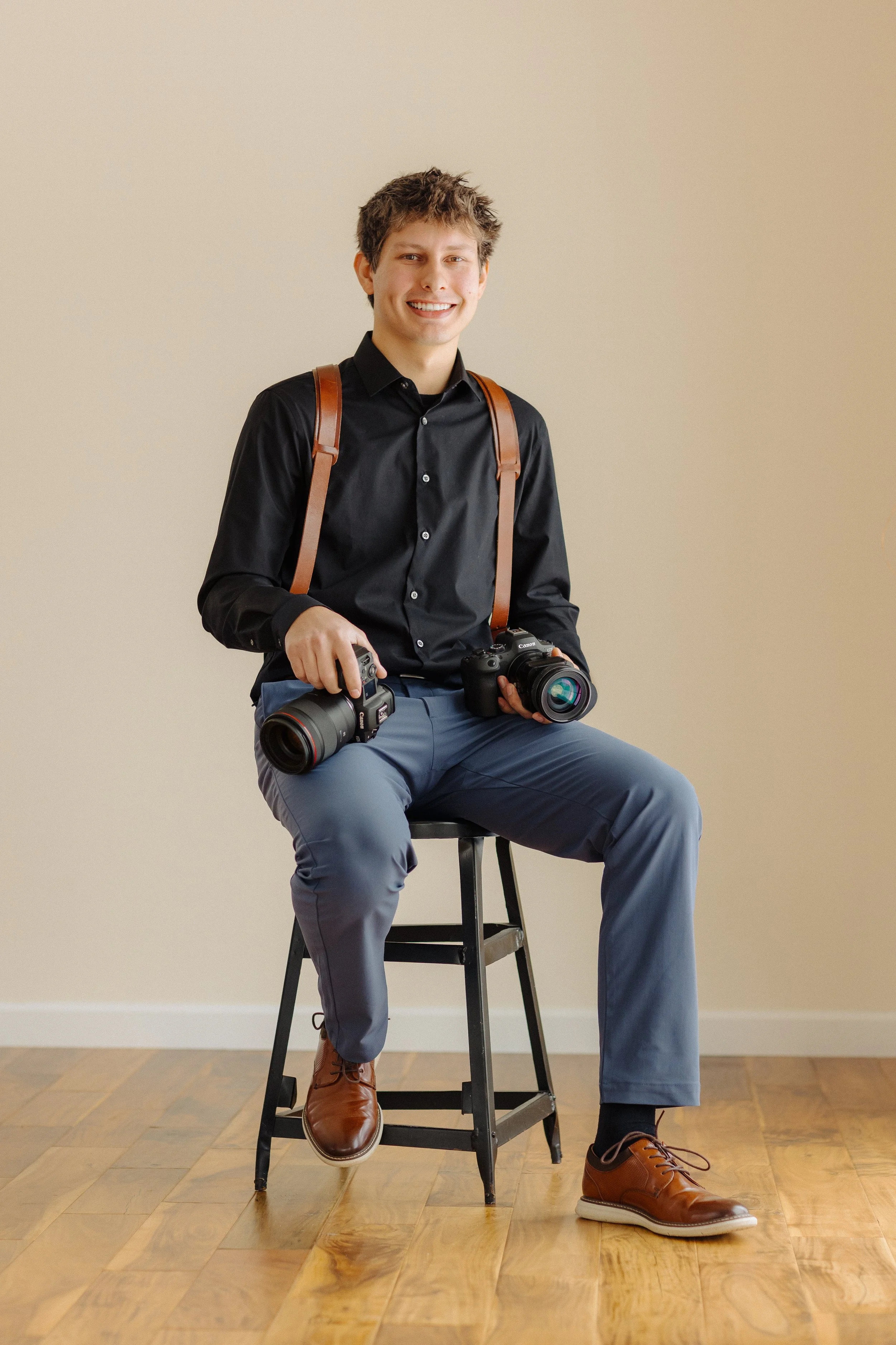 A young man with brown hair, wearing a black shirt, gray pants, and brown shoes, sitting on a black chair against a plain beige wall. He is smiling and holding two professional cameras with lenses, with camera straps over his shoulders.