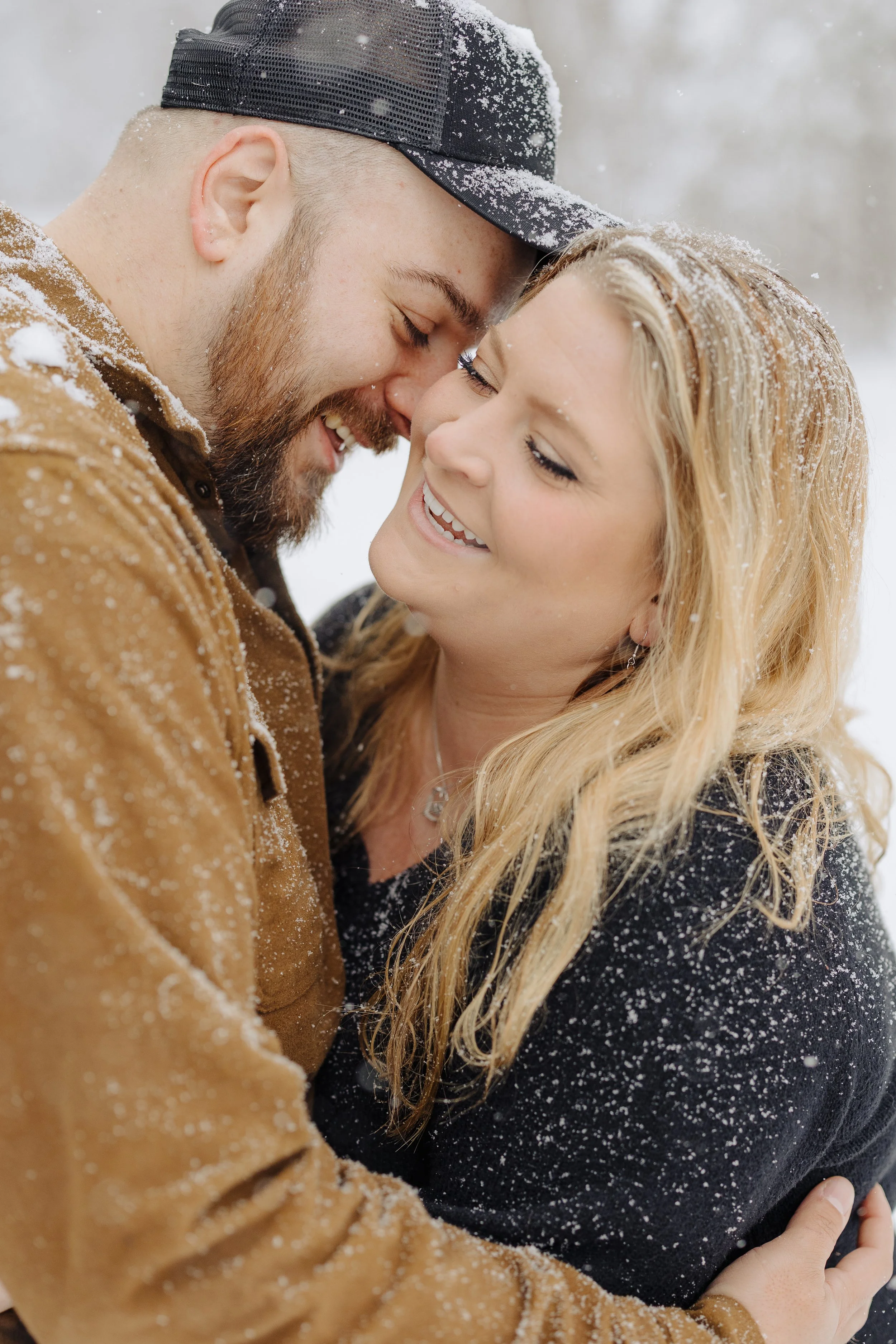 A smiling couple embracing outdoors in snowy weather, with their foreheads touching and eyes closed.