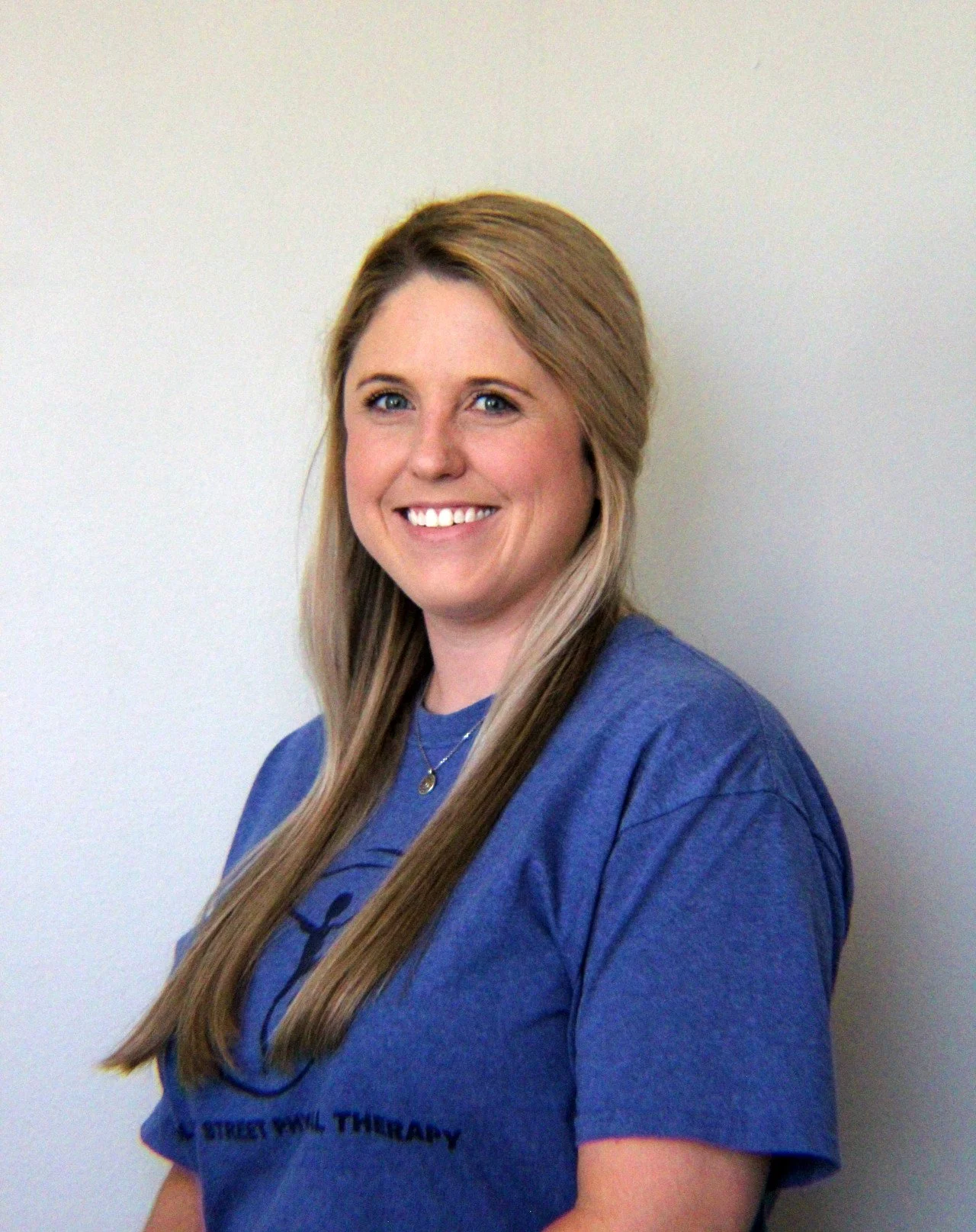 Young woman with long blonde hair smiling, wearing a blue T-shirt with text 'Street-Ville Therapy,' against a plain light-colored background.