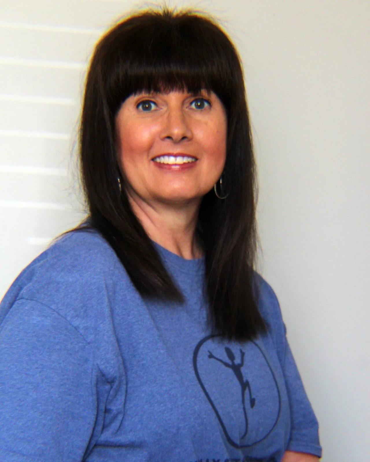 A woman with long dark hair and bangs, wearing a blue t-shirt with a logo, smiling and standing in front of a plain wall.