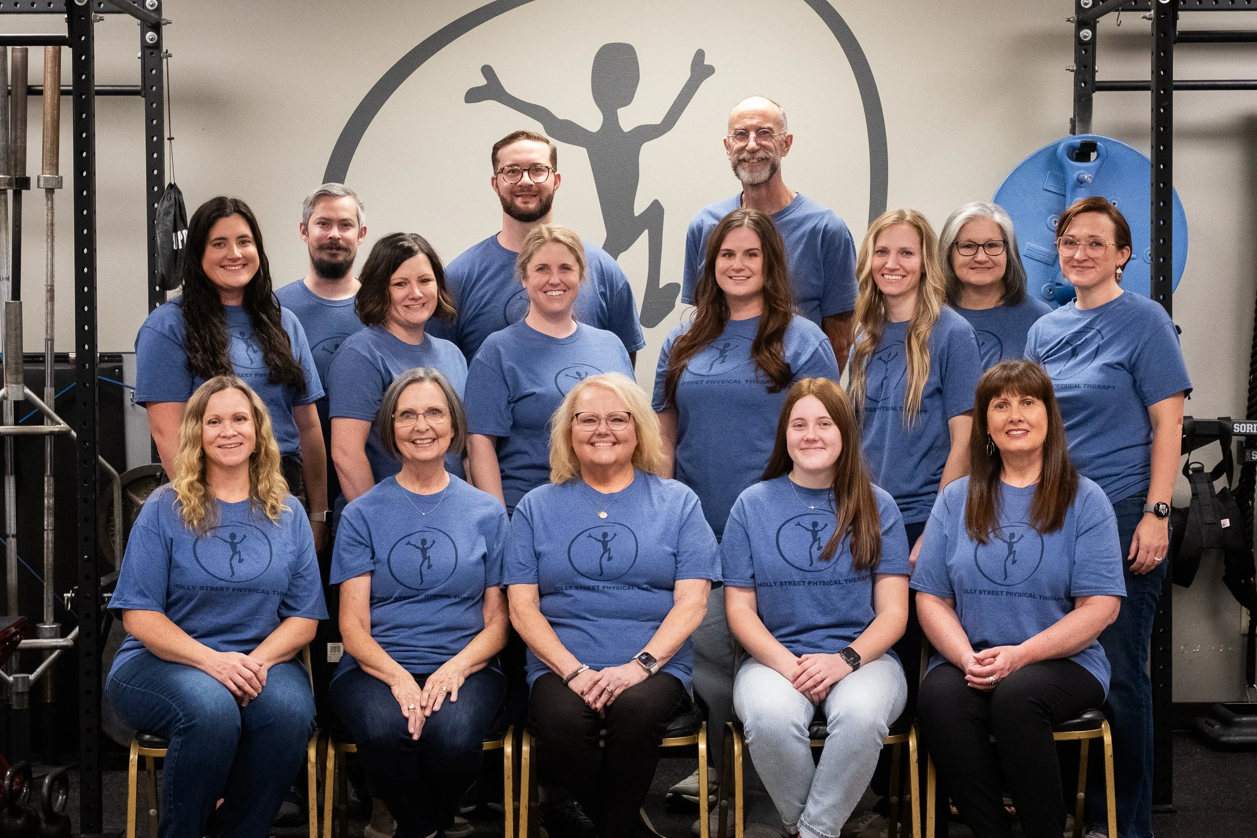 Group photo of 14 people, mostly women, in matching blue t-shirts with a logo of a person dancing inside a circle, posing in a gym or therapy center with fitness equipment in the background.