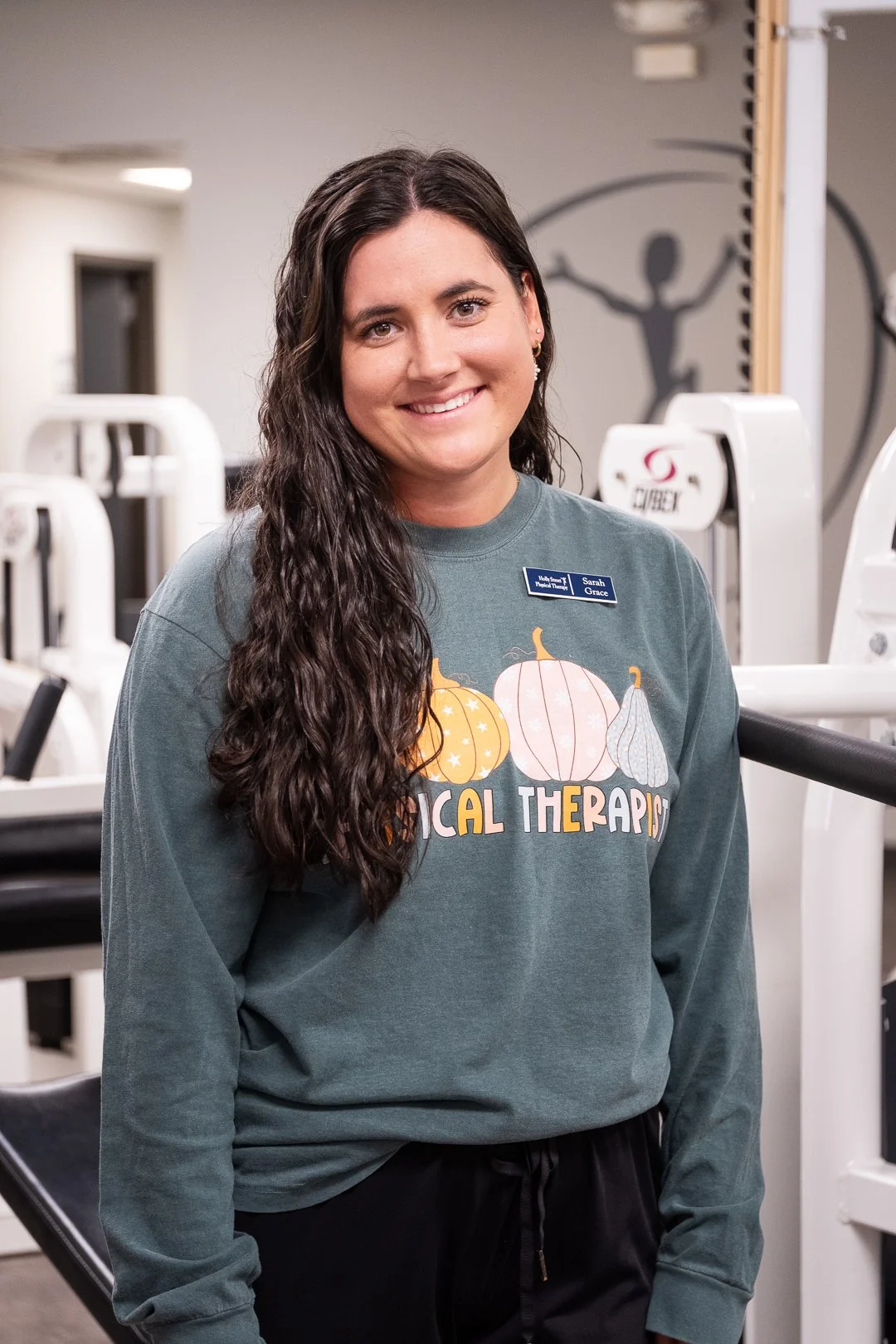 A woman with long wavy dark hair smiling in a gym. She is wearing a gray sweatshirt with pumpkins and the words 'Natural Therapies' and a name tag that says 'Sarah Grace.'