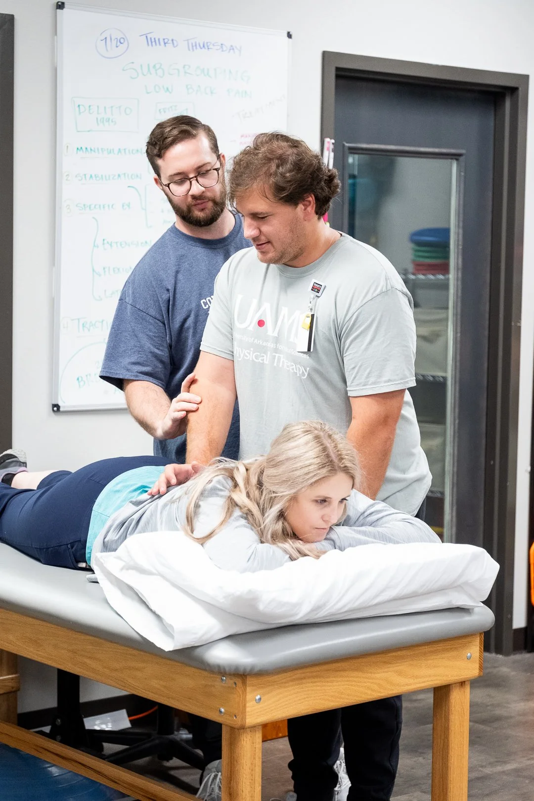 A physical therapy session with a woman lying on a treatment table, two men assisting her, and a whiteboard with notes in the background.