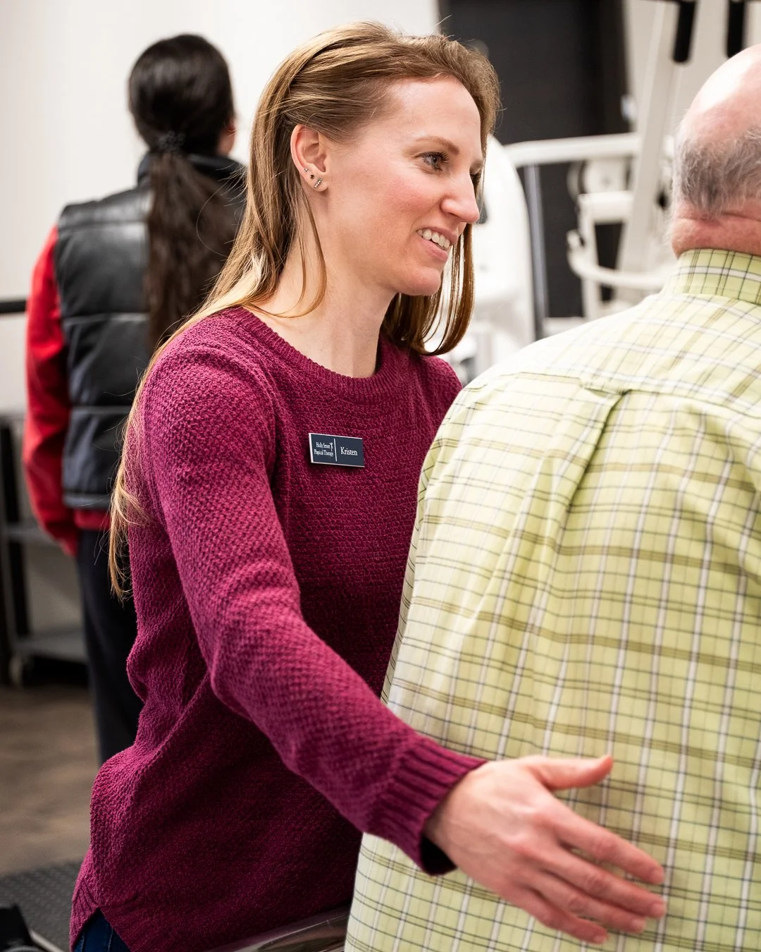 A woman wearing a maroon sweater with a name tag, smiling and talking to a man in a yellow plaid shirt in an indoor setting.