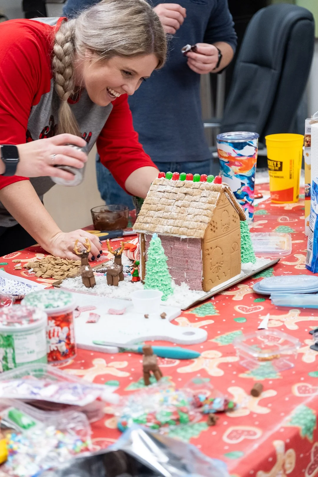 A woman decorating a gingerbread house at a holiday party, with a table filled with festive treats and decorating supplies.