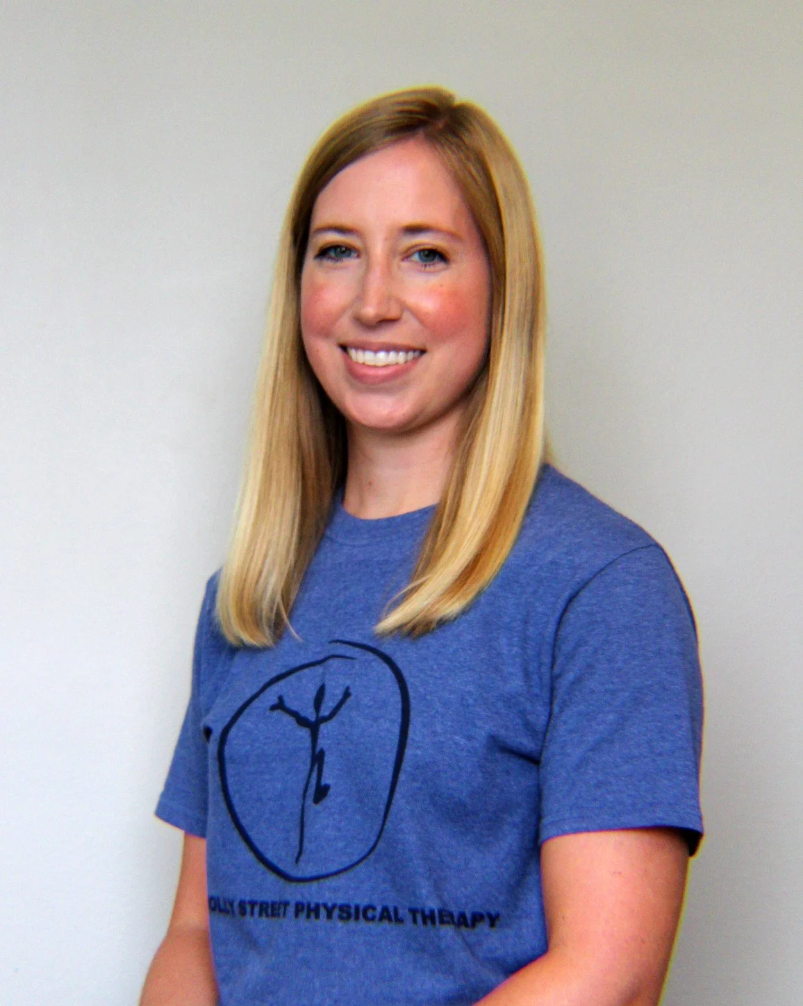 A woman with blonde hair smiling, wearing a blue T-shirt with a logo and text for Hollywood Street Physical Therapy, standing against a plain light-colored background.