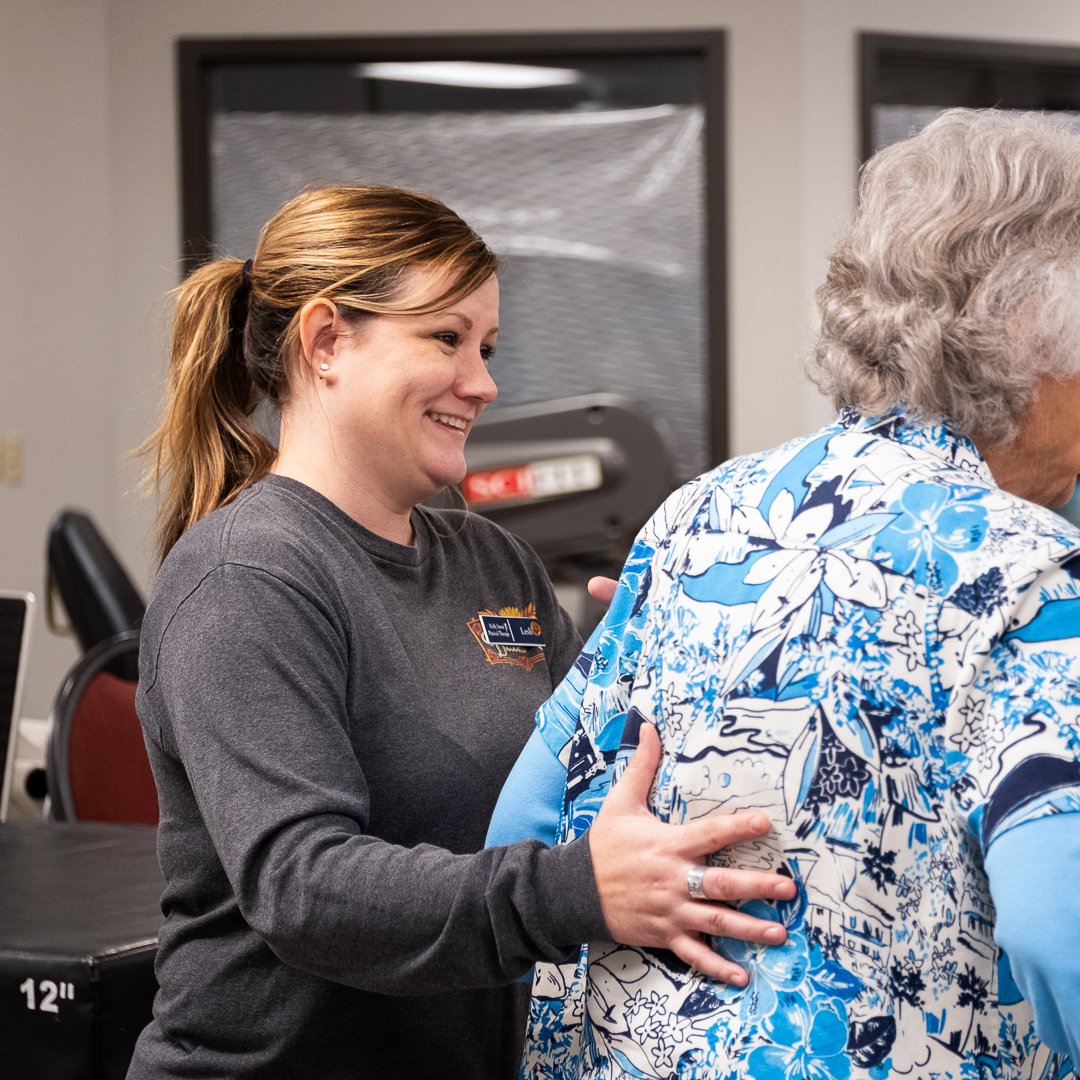A woman with brown hair tied back, smiling, assisting an elderly person with gray hair, dressed in a blue and white floral shirt, in an indoor setting.