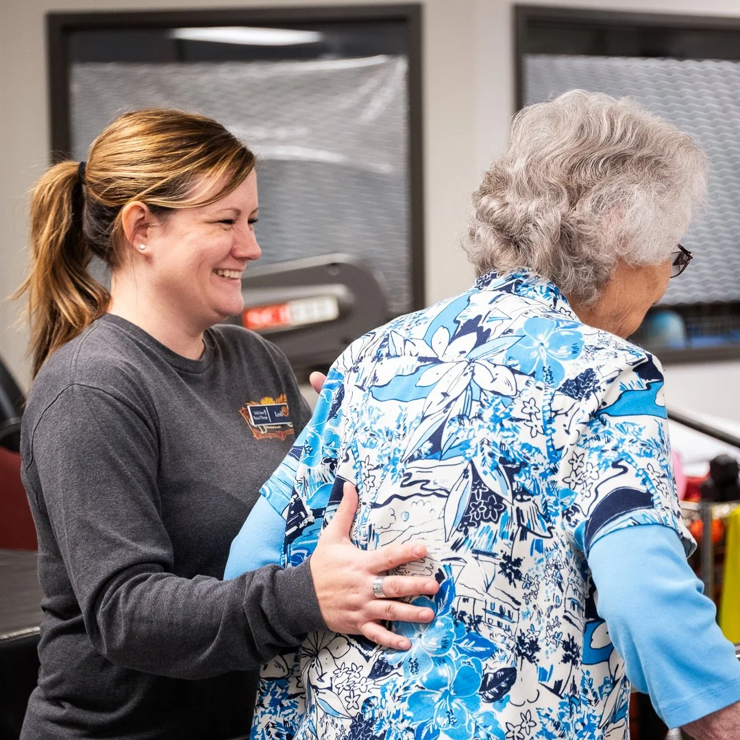 A young woman with a ponytail and wearing a gray shirt is smiling and gently touching an elderly woman with gray curly hair and glasses, who is wearing a blue and white floral-patterned shirt.