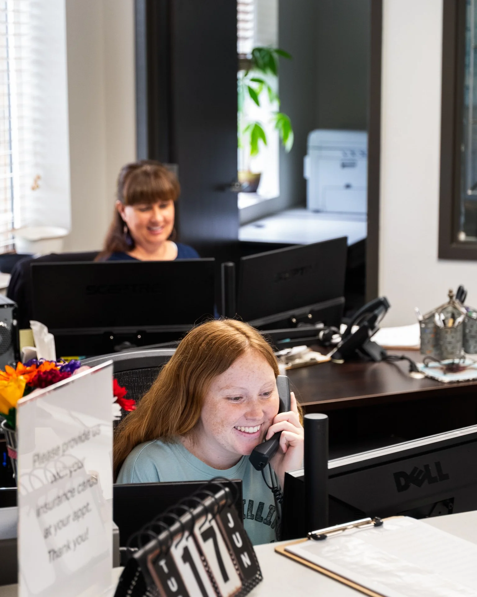Two women working at desks in an office, one on the phone smiling, with computers and office supplies around.
