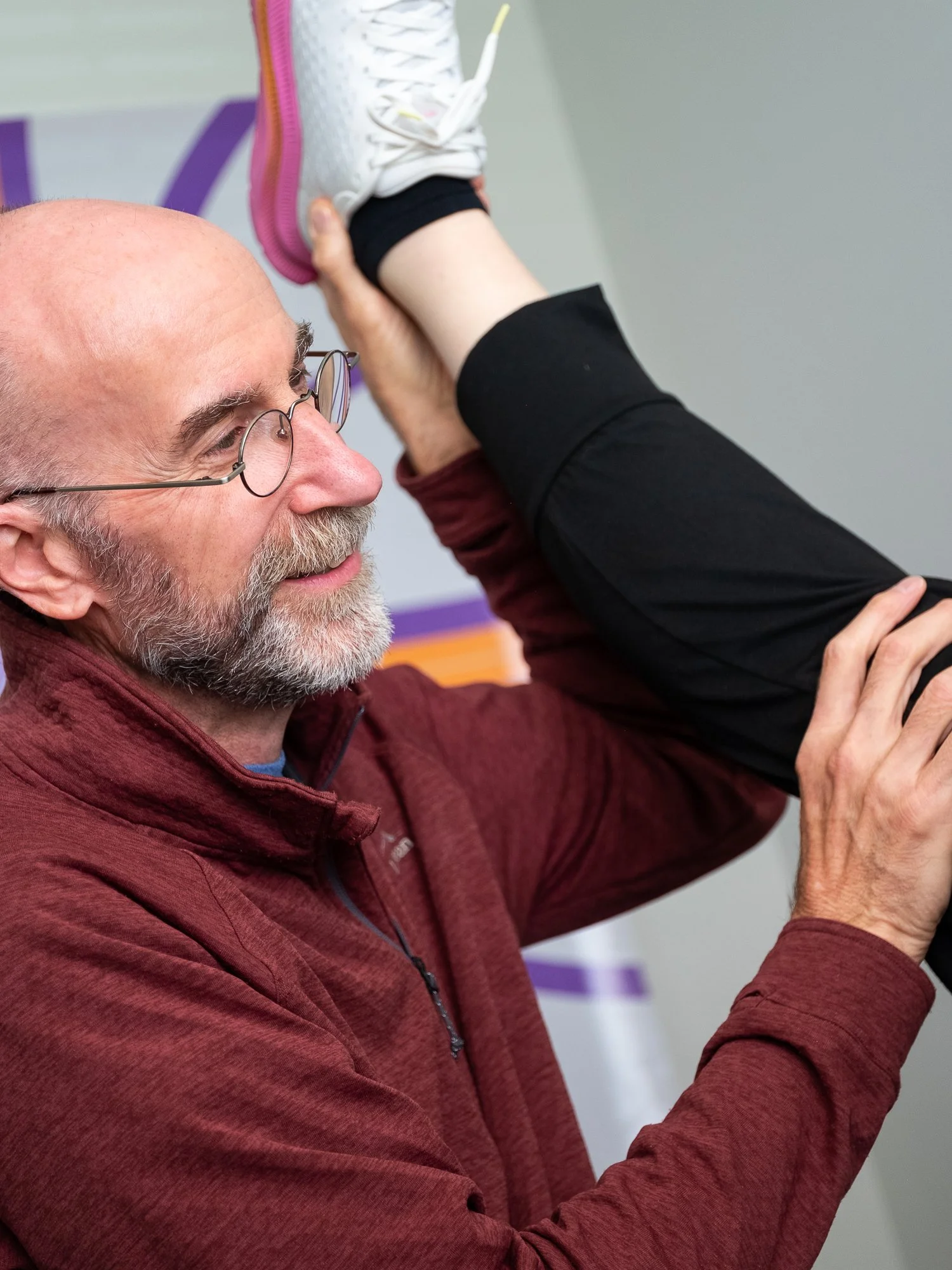 An older man with glasses, gray beard, and maroon jacket helps a person stretch by pulling their leg up behind their head during a fitness session.