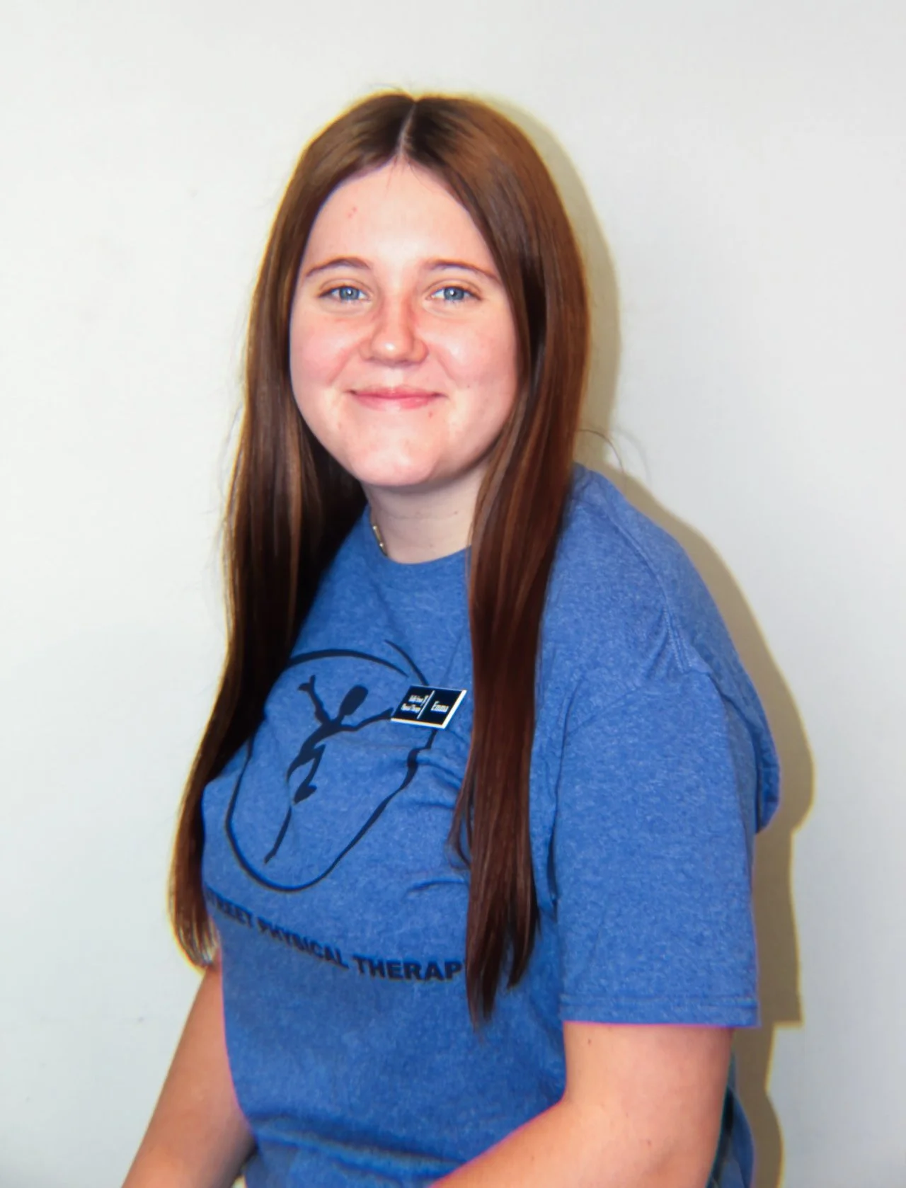 A young woman with long red hair, blue eyes, wearing a blue t-shirt with a logo and text for physical therapy, smiling, standing against a white wall.