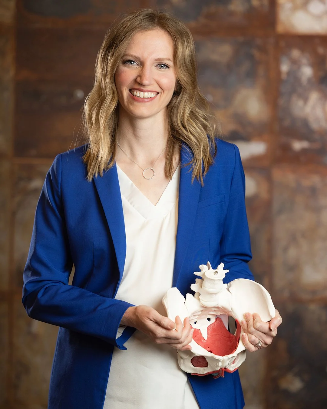 Woman with light brown hair smiling and wearing a blue blazer holding a human pelvis model with red and white internal structures, in front of a blurred brick wall background.
