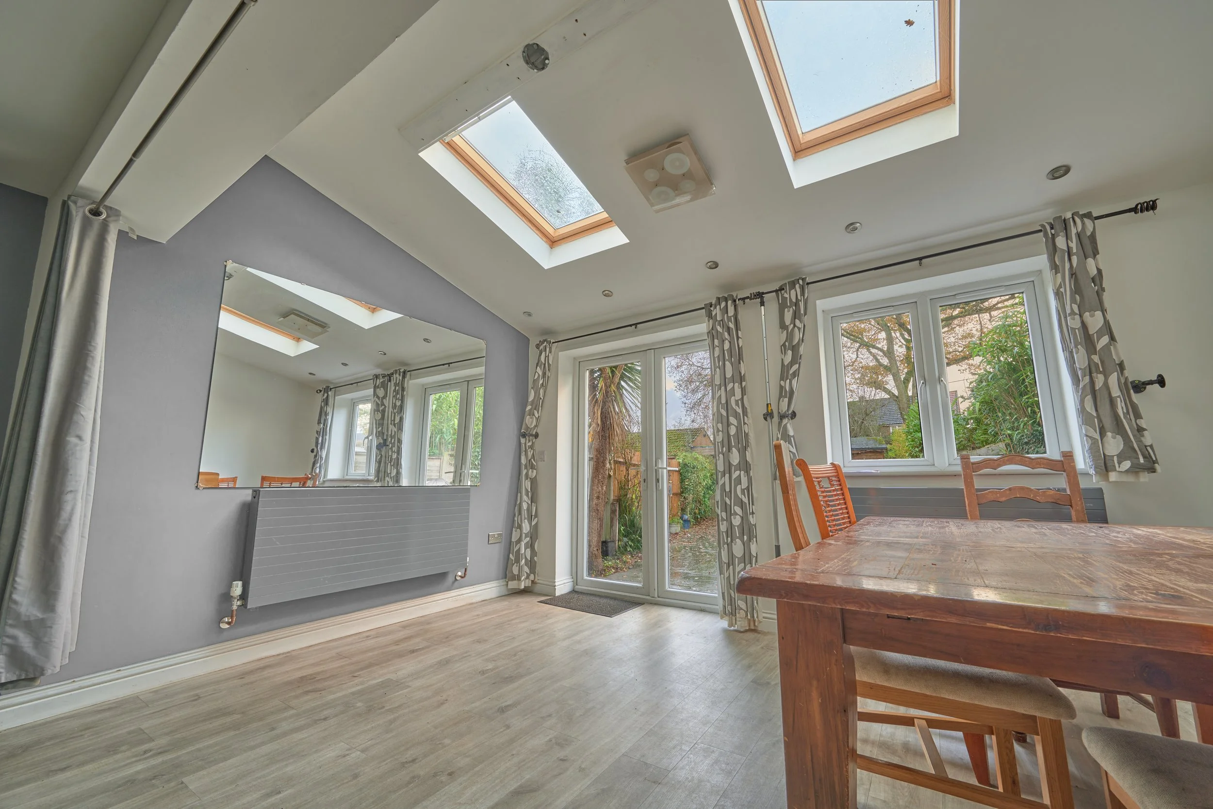 Dining area with large windows and skylights, wooden table and chairs, curtain rods, and a wall mirror reflecting part of the room.