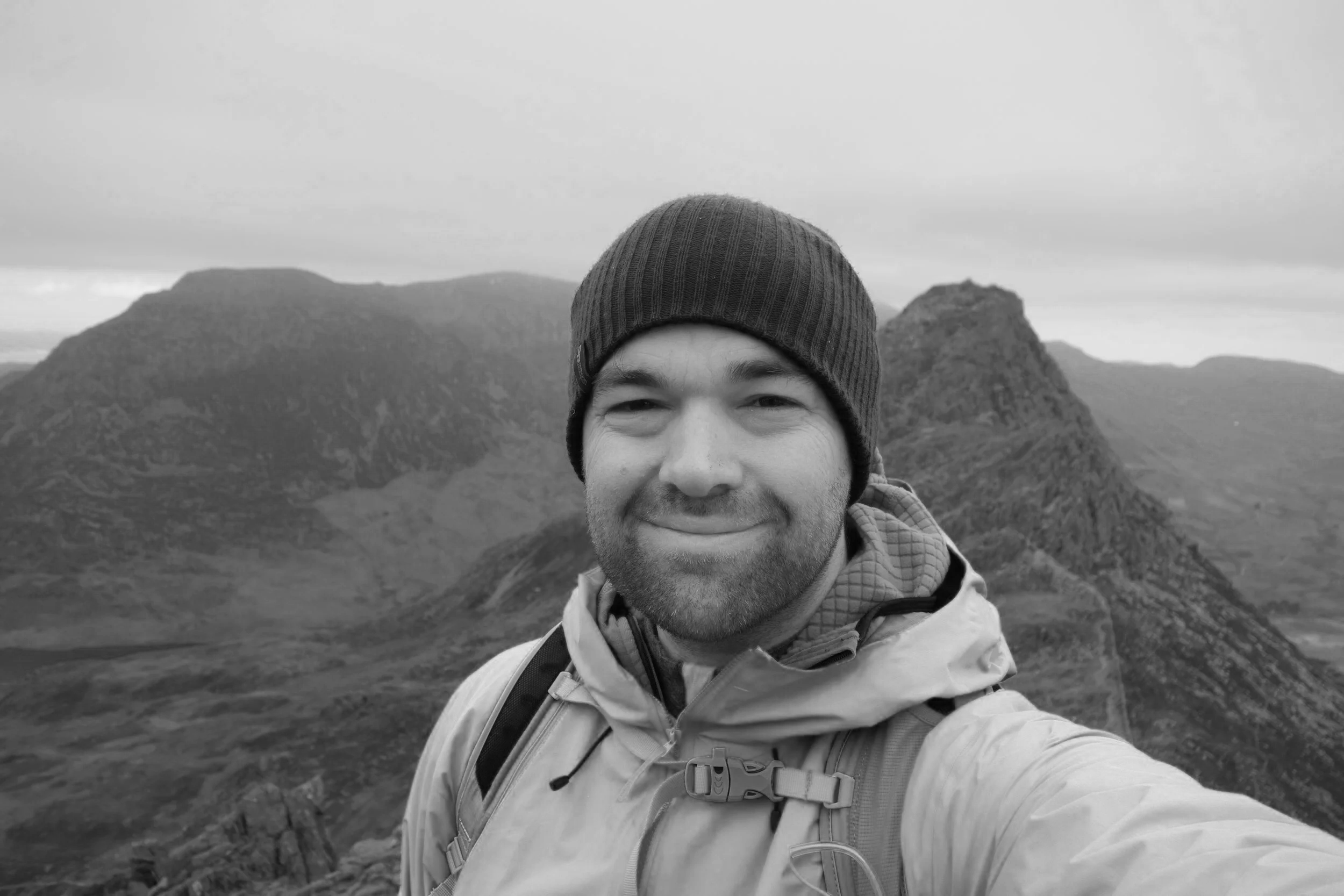 A man smiling in outdoor gear, wearing a beanie and jacket, taking a selfie with a mountainous landscape in the background.