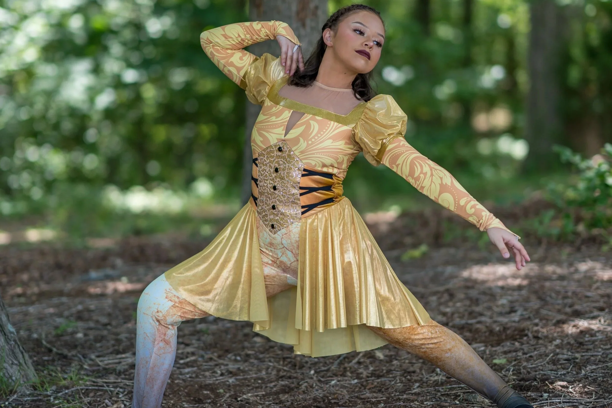 Crystal Najera dressed in a yellow costume with brown and gold accents poses outdoors among trees, with one arm raised and one arm extended to the side.