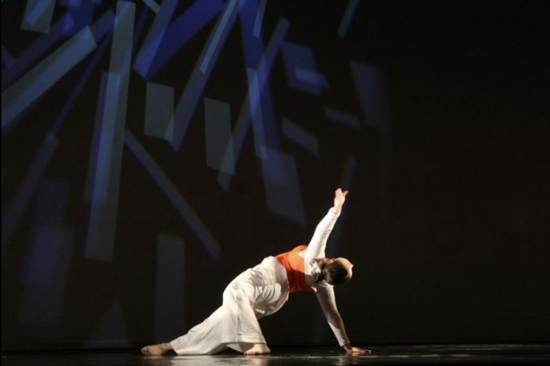 Crystal Najera performing a dance move on stage, dressed in white pants and an orange top, with one hand on the ground and the other raised in the air, against a dark background with blue geometric patterns.