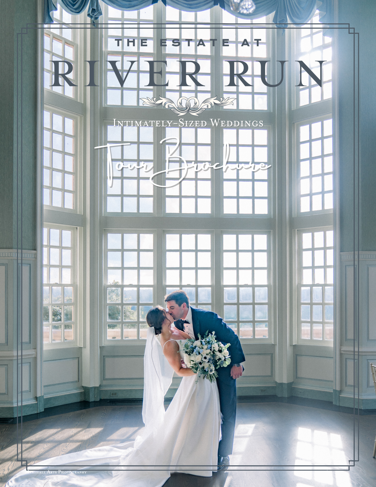 A wedding couple standing in front of a large windowed wall, sharing a kiss. The bride is holding a bouquet of white and blue flowers, and the groom is dressed in a tuxedo. Bright natural light filters through the windows, illuminating the scene.