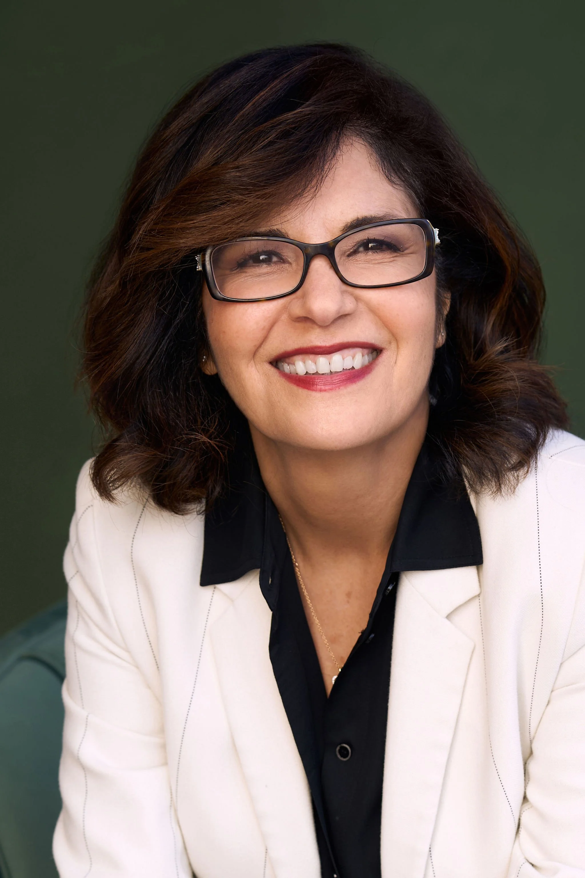 A woman with dark brown hair, glasses, and a white blazer smiling at the camera against a green background.