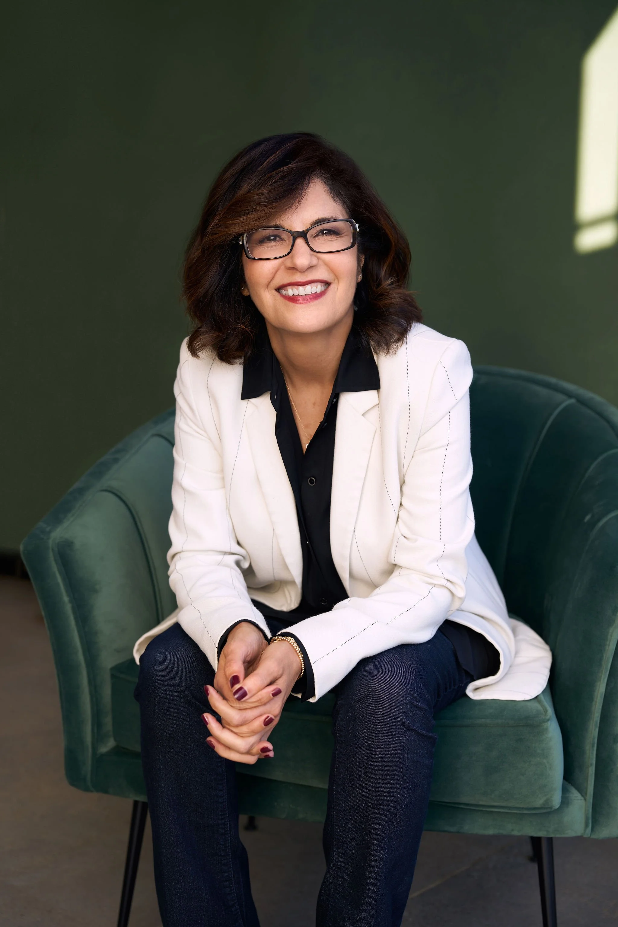 Smiling woman with dark hair, glasses, wearing a white blazer over a black shirt, sitting on a green chair against a dark green background.