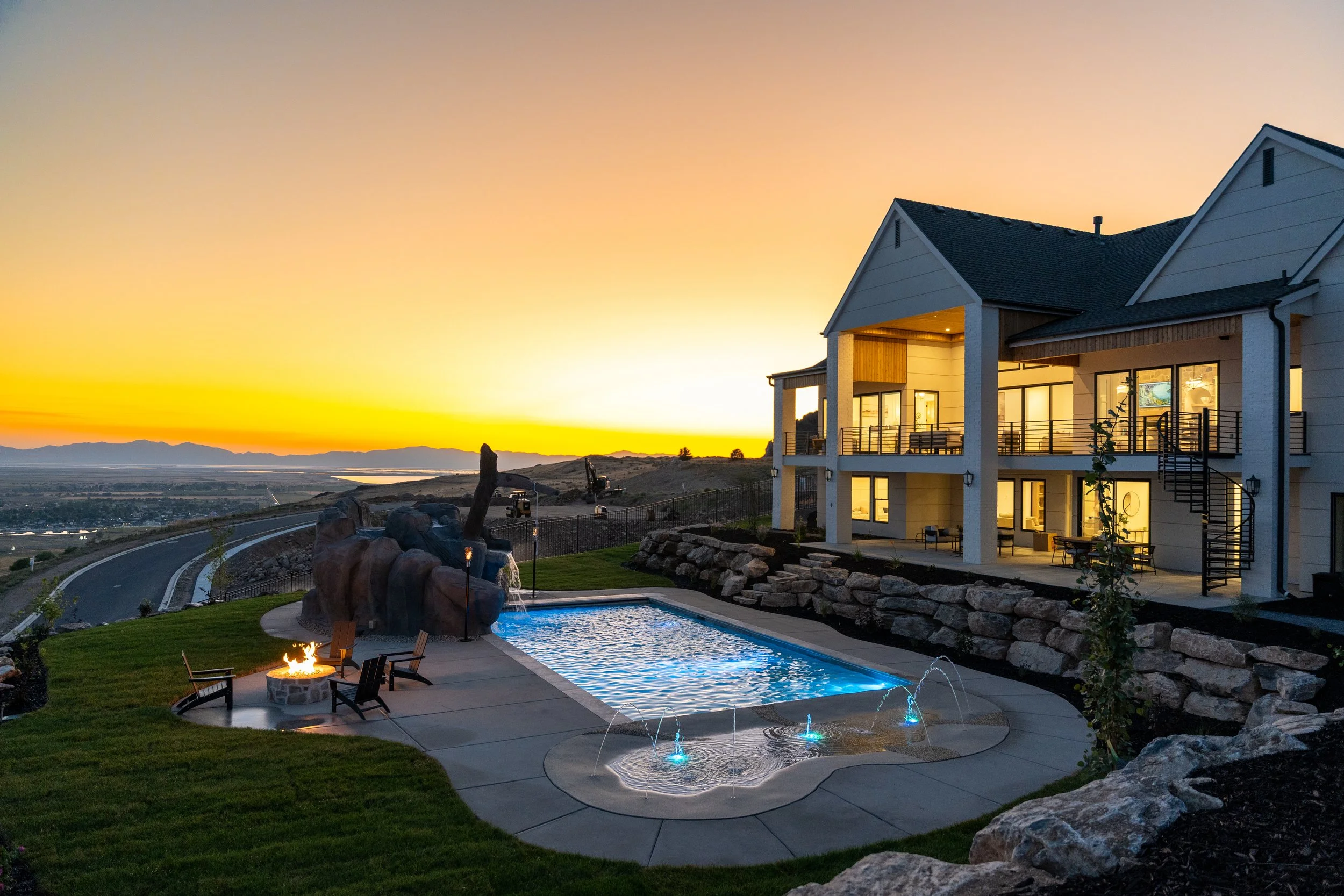 A modern house with multiple stories and large windows overlooking a scenic landscape at sunset. In the foreground, there is a swimming pool with a fountain feature, a fire pit with chairs around it, and a landscaped yard with rocks and a small tree. The house has outdoor balconies and staircases.