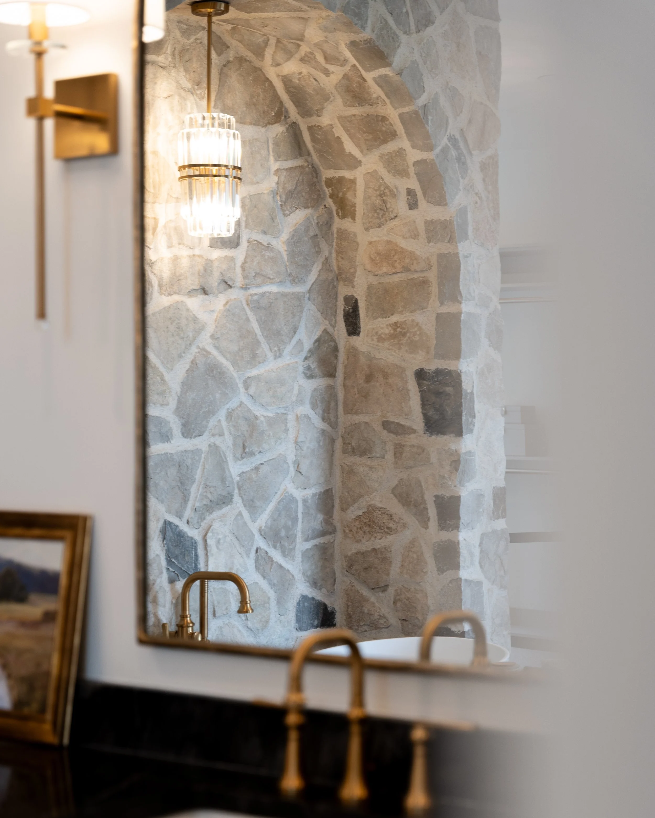 A bathroom with a stone wall, a pendant light, and a gold faucet reflected in a mirror.