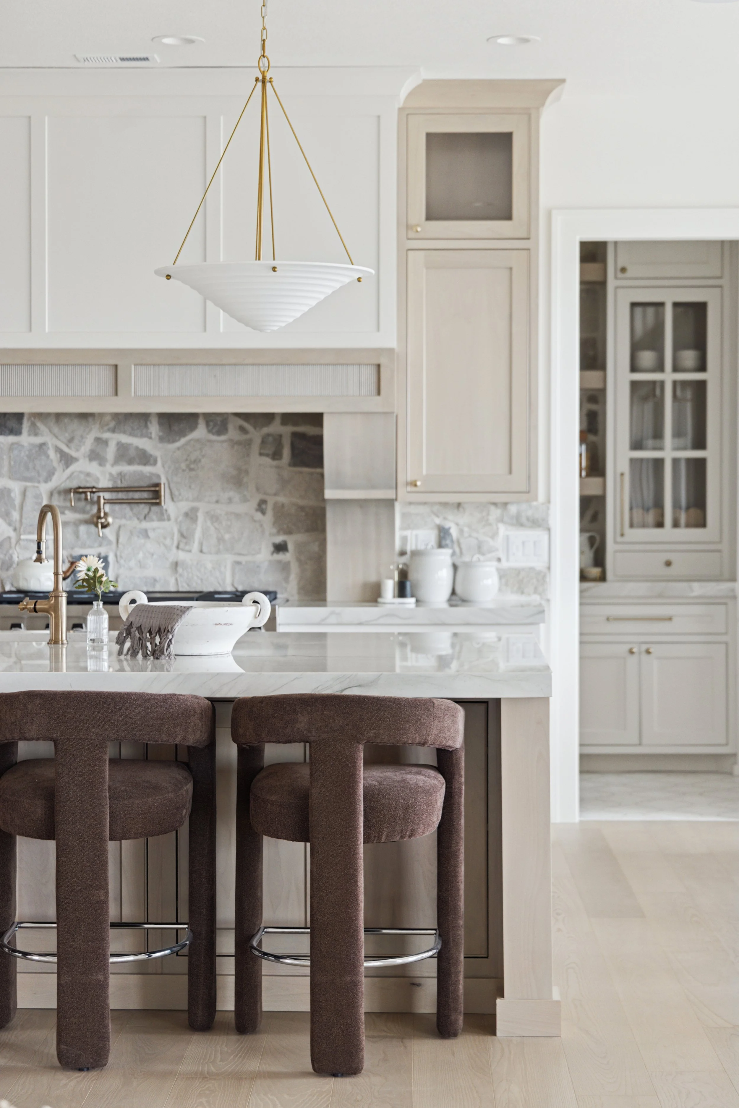 Modern kitchen with white cabinets, stone backsplash, marble countertop, brown upholstered bar stools, gold pendant light, and decorative white ceramics.