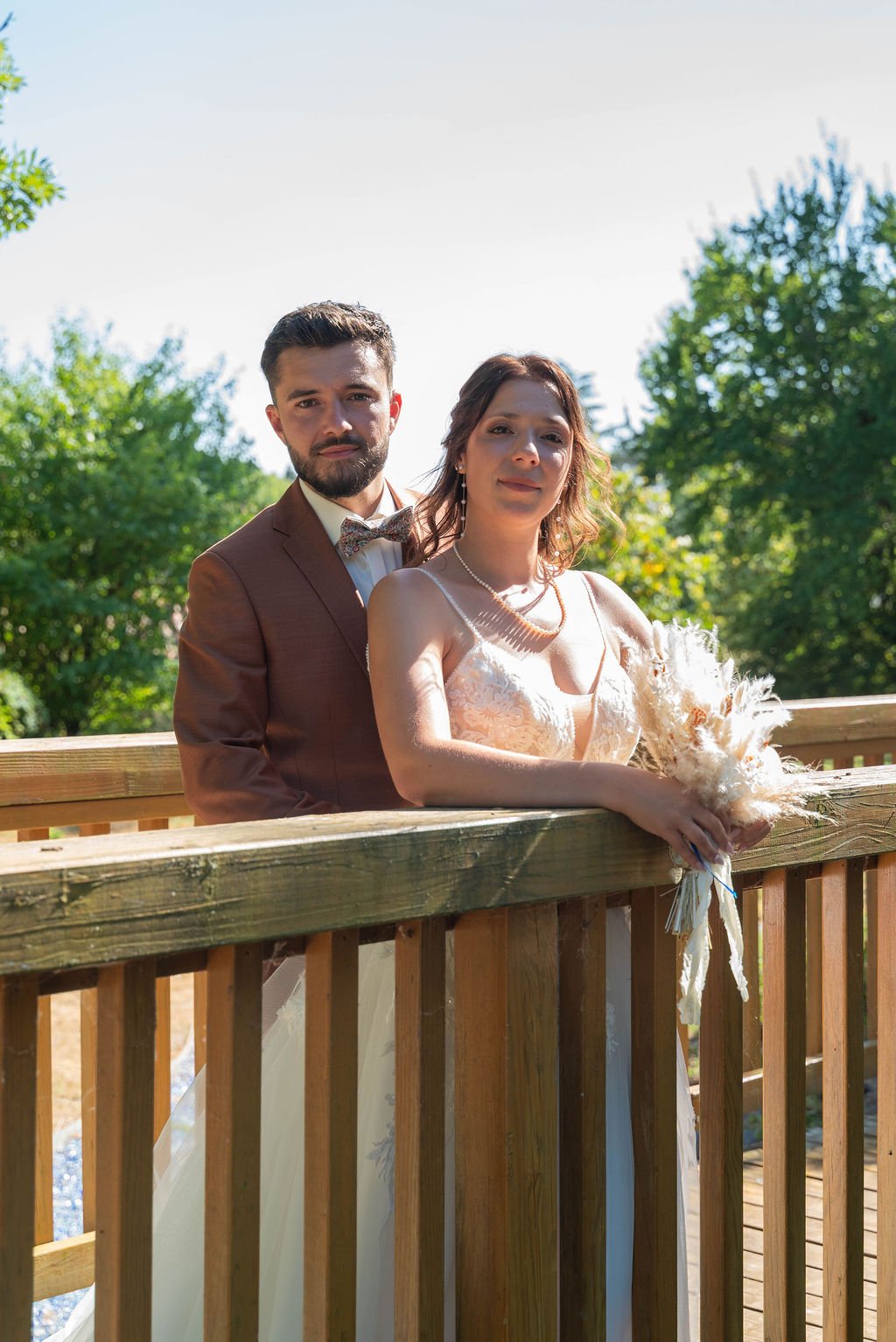 Un couple en vêtements de mariage se tient sur une terrasse en bois entourée d'arbres verts, sous un ciel clair.